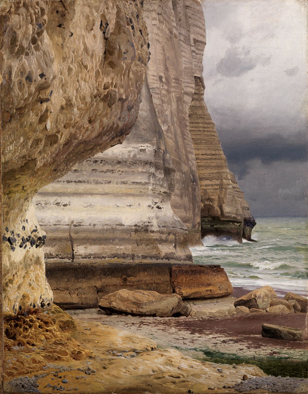 Schirmer zeigt eine beeindruckende Küstenlandschaft mit massive Felsen, die sich steil aus dem Meer erheben, umgeben von schäumenden Wellen. Der Himmel ist teils bewölkt.