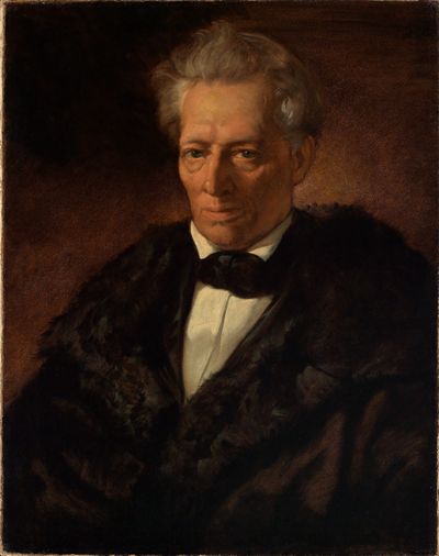 Das Bild zeigt Professor Theodor Welcker in einer ruhigen und nachdenklichen Pose. Er trägt einen dunklen Anzug und sitzt auf einem Stuhl. Sein Gesicht wirkt ernst und konzentriert.