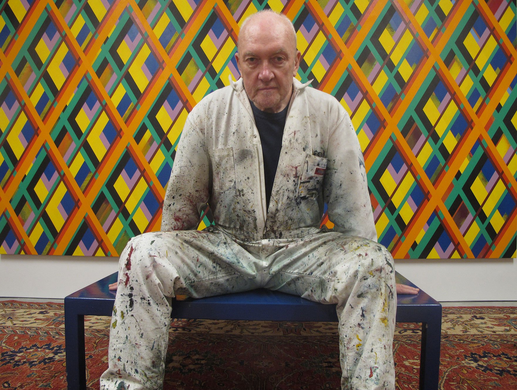 Der zeitgenössische Künstler Sean Scully sitzt ein einem mit Farbe beschmutzen Maler-Overall auf einer Bank vor seinem Werk Overlay 2. DasWerk zeigt diagonale bunte Linien, die zusammen eine Vielzahl von Rauten bilden.
