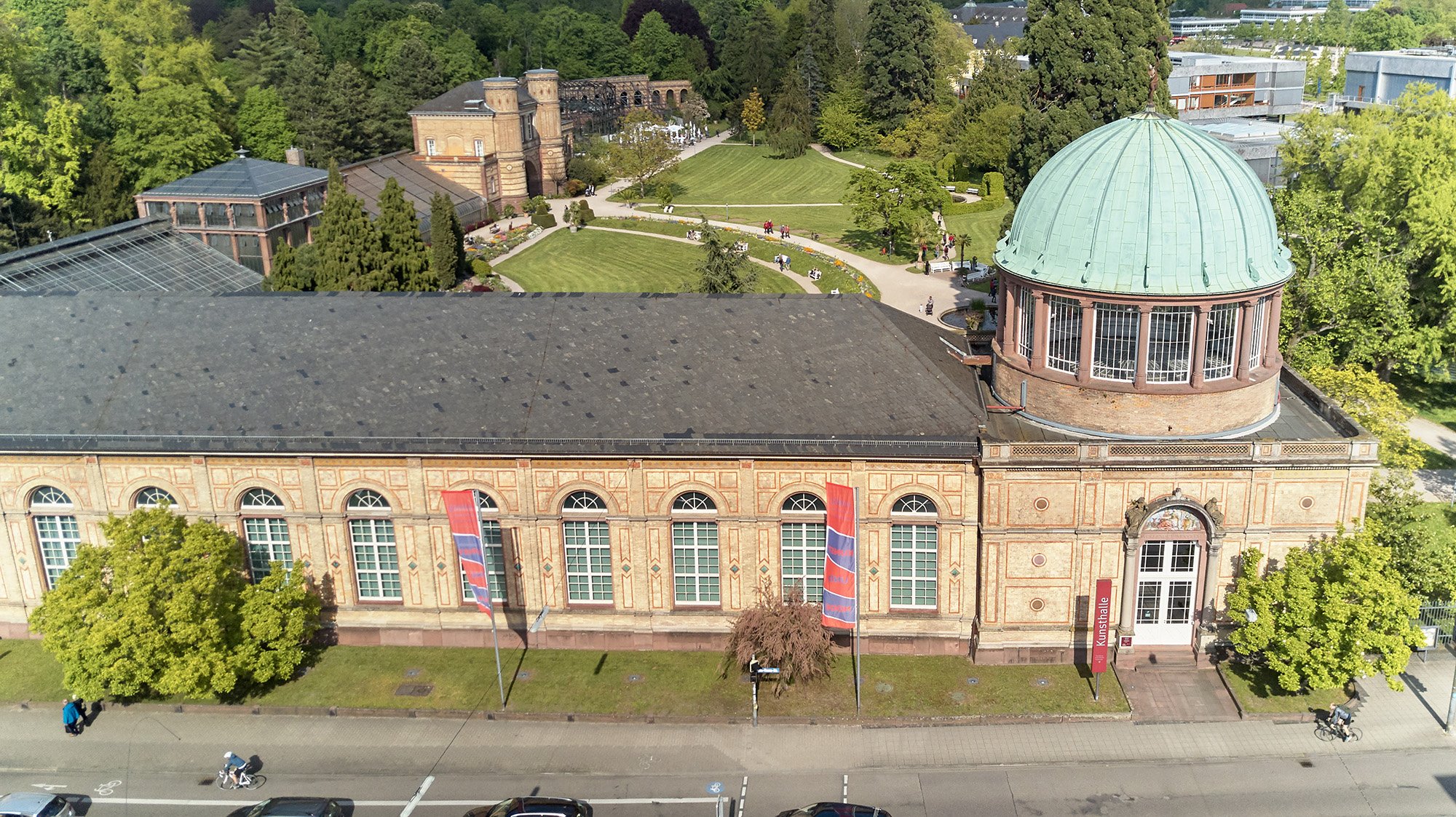 Foto der Orangerie der Staatlichen Kunsthalle Karlsruhe. Der Bau ist lang und hat hohe Rundbogenfenster. Am rechten Ende es Gebäudes hat es eine Kuppel.