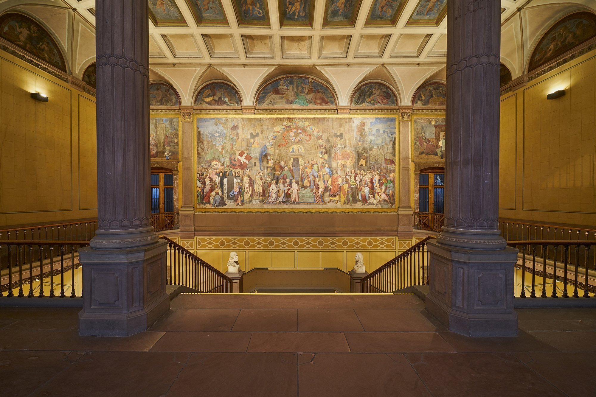 Foto des Treppenaufgangs im Hauptgebäuder der Kunsthalle Karlsruhe mit dem Schwind-Fresko. Das Fresko zeigt eine große Personengruppe, die auf ein Kirchenportal zugeht.
