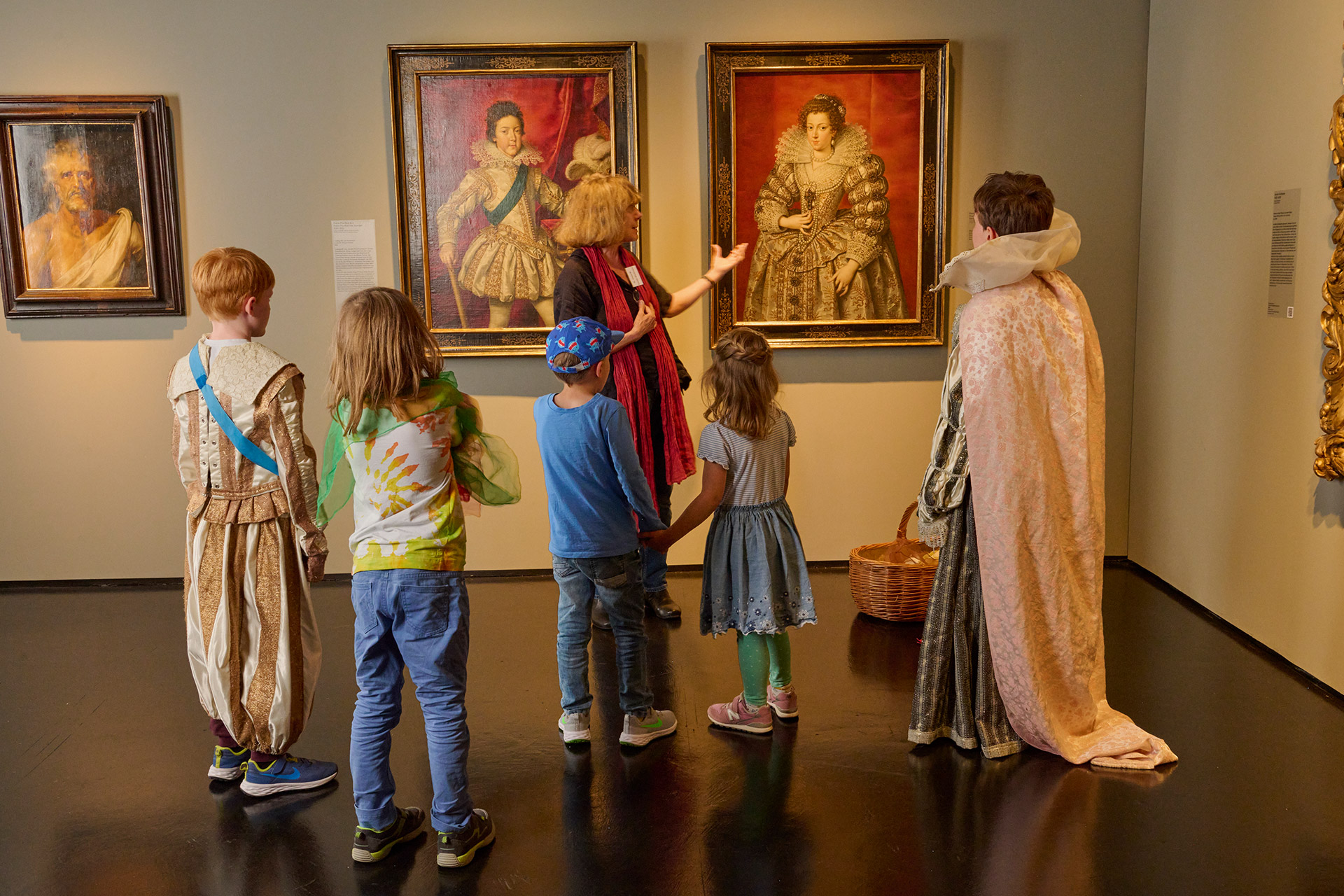 Mehrere Kinder stehen verkleidet in der Ausstellung und betrachten zwei Gemälde, auf denen die Porträtierte ähnlich prunkvolle Kleidung tragen.
