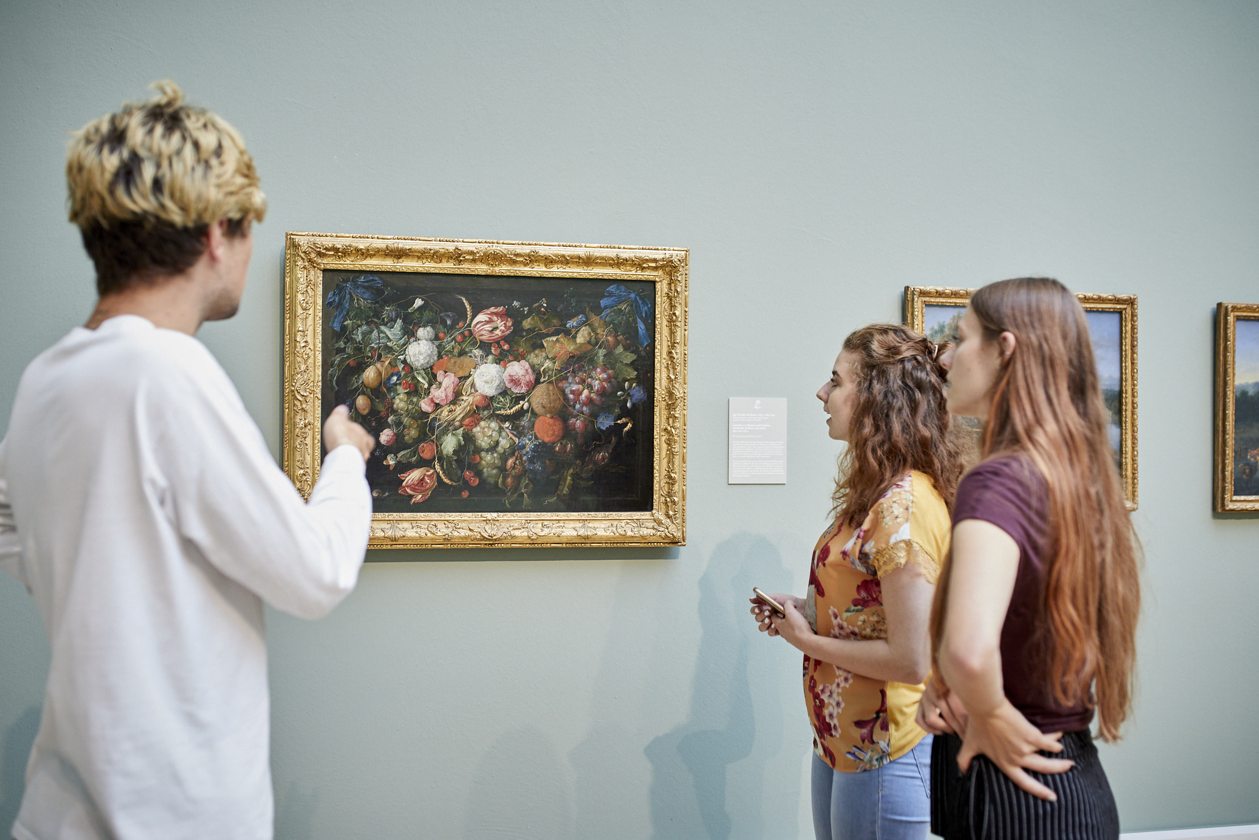 Blick in die Ausstellung, in der sich junge Personen umschauen und ein Blumenstillleben betrachten.