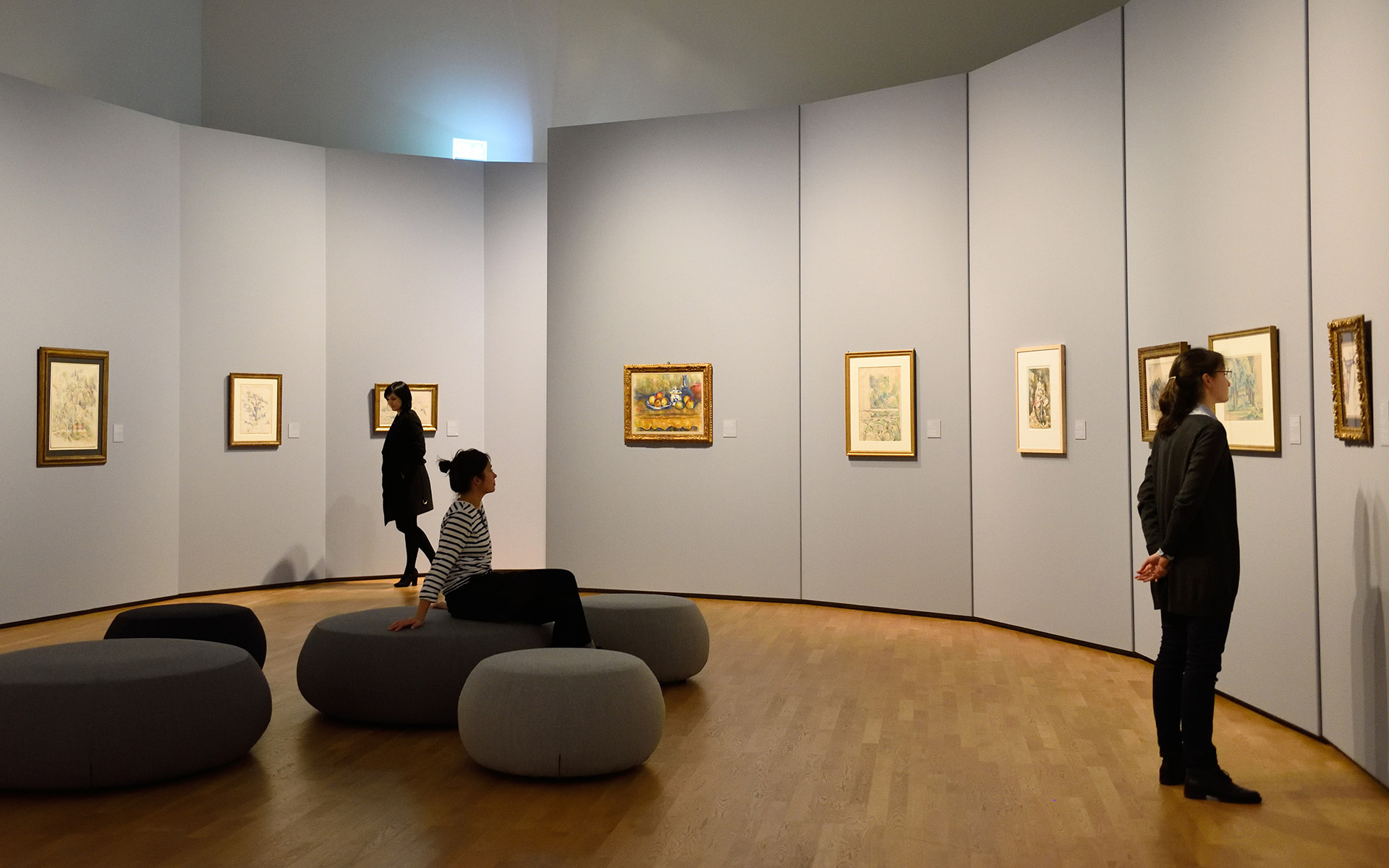 Ein ovaler Raum mit Sitzgelegenheiten in der Mitte. An den Wänden hängen Bilder die von drei Personen betrachtet werden.