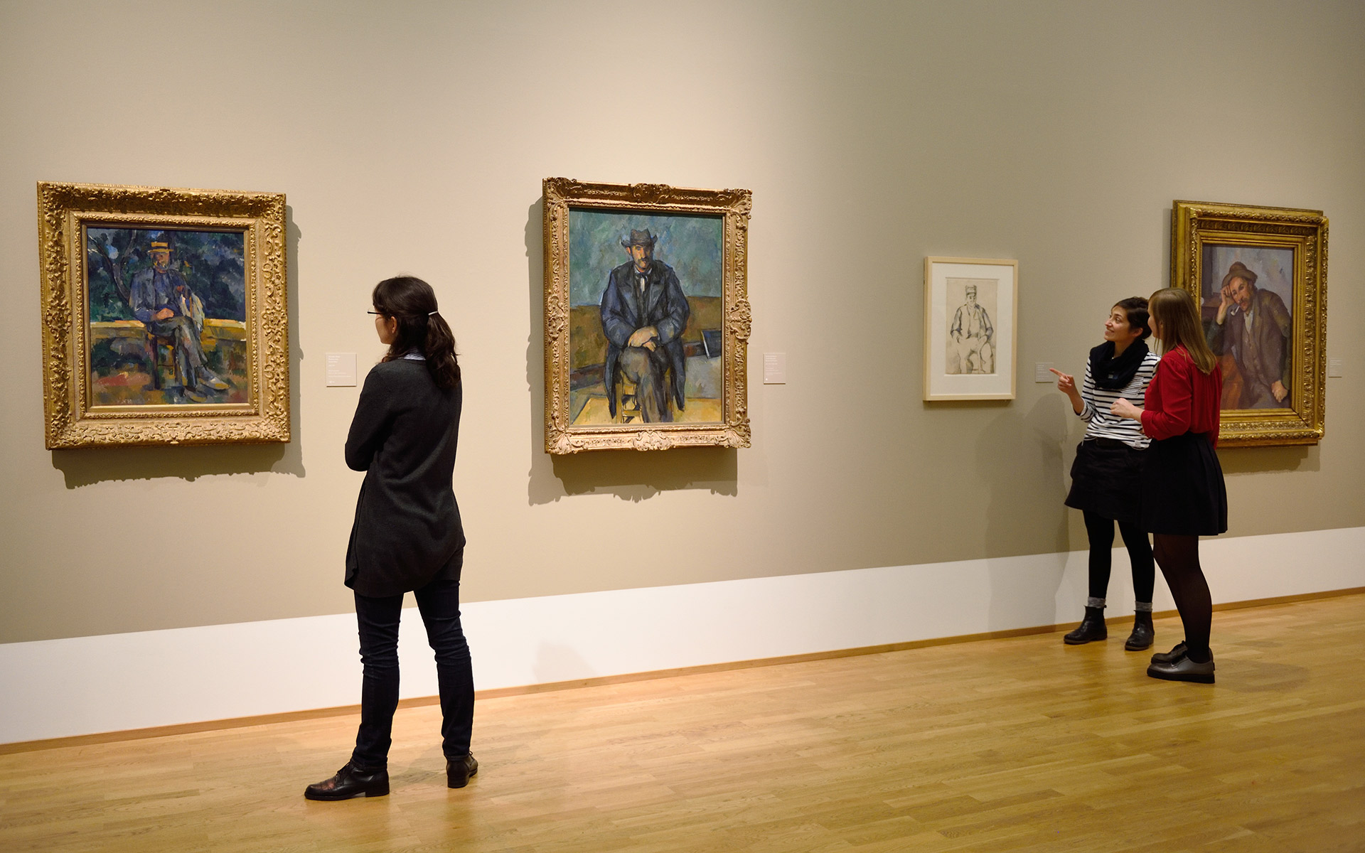 Ein Museumsraum mit vier Gemälden und drei Personen, die diese betrachten.