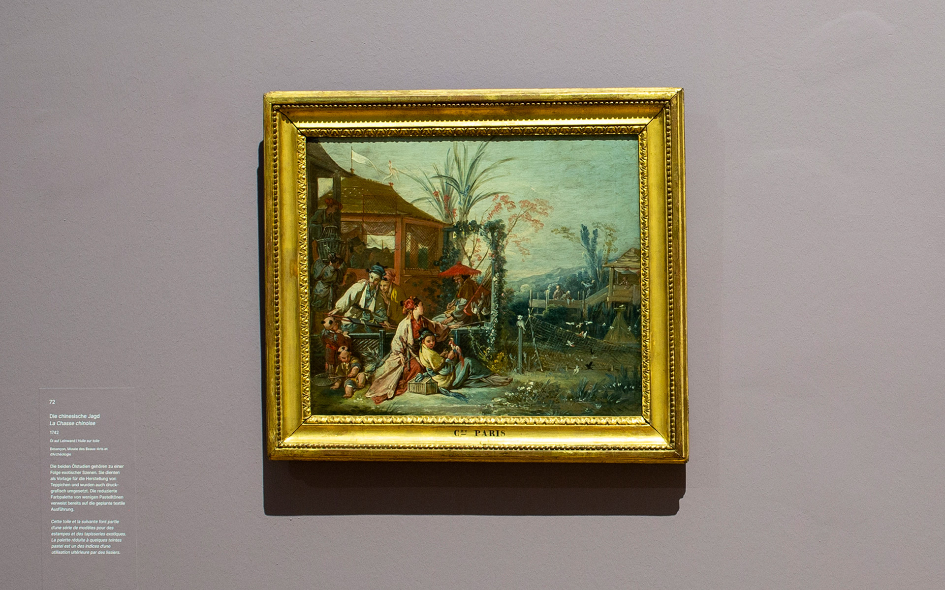 Ausstellungsansicht des Gemäldes Die chinesische Jagd von 1742 des Künstlers François Boucher. Das Gemälde hat einen Goldrahmen. Links daneben befindet sich ein Text zu dem Bild auf der Wand.
