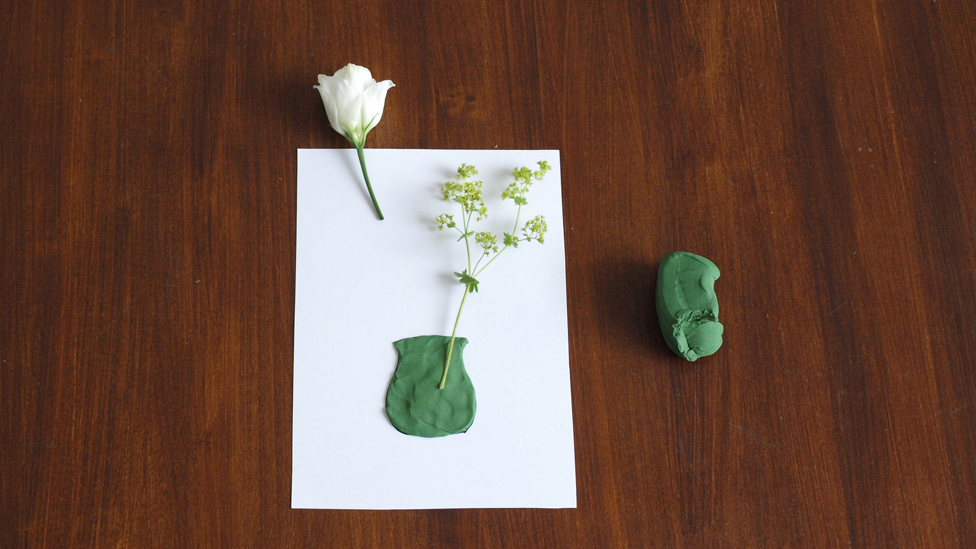 Auf einem Tisch liegt ein Blatt Papier mit einer Knetvase und zwei Blumen.