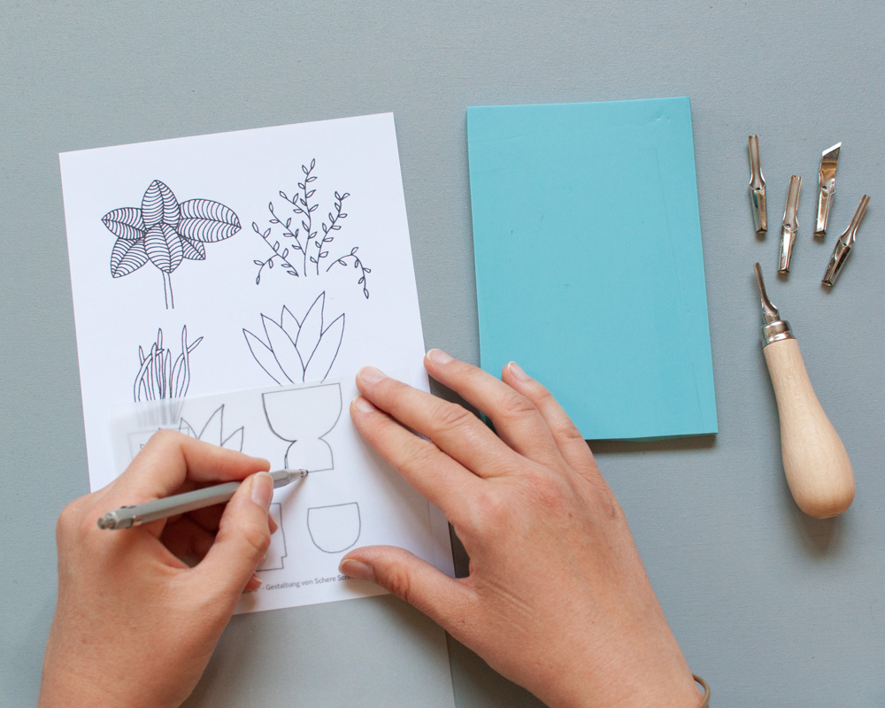 Zwei Hände zeichnen auf einem Blatt Papier. Daneben liegt ein Werkzeug und blaue Pappe.