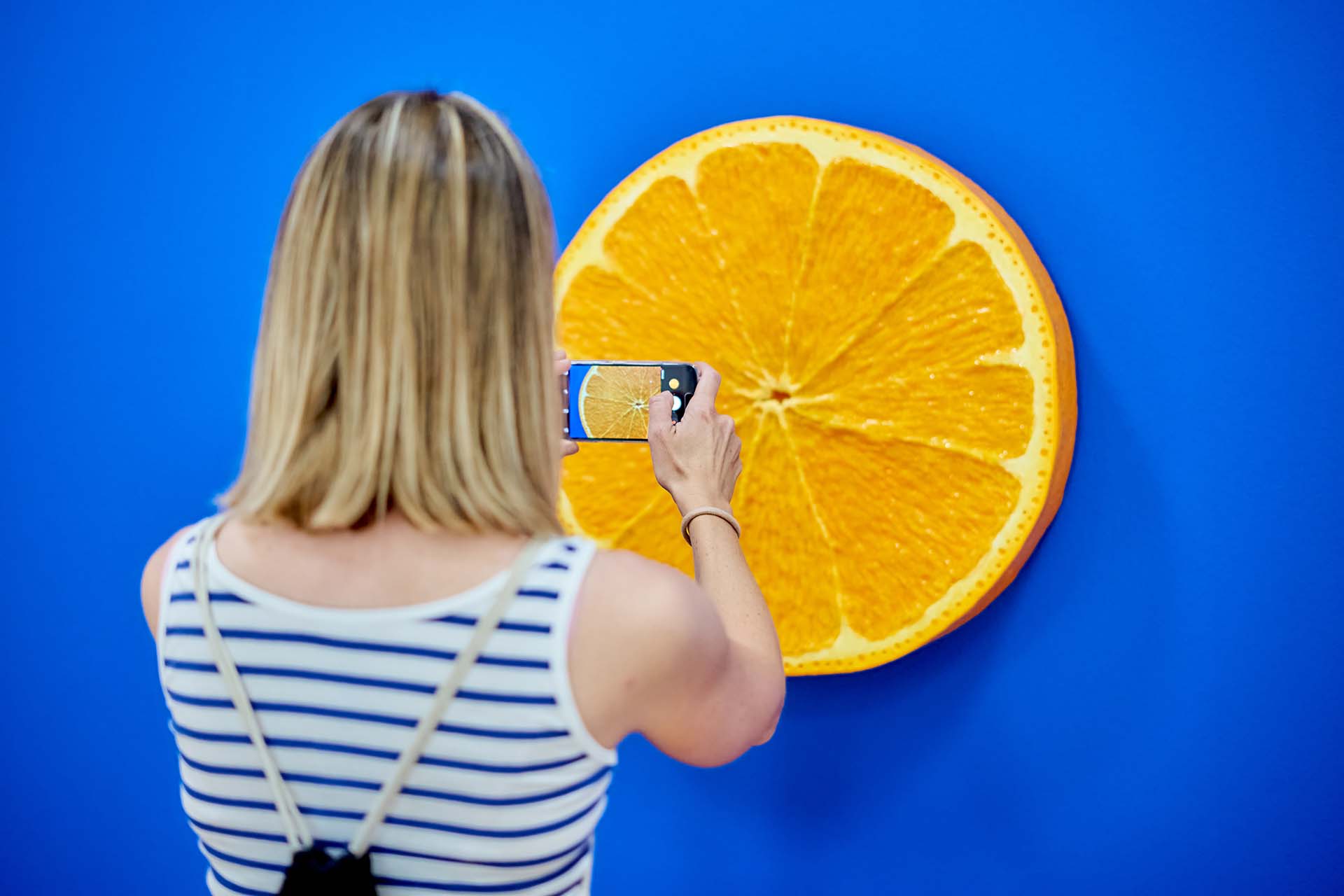 Frau, die von hinten beim Fotografieren eines orangenscheibenförmigen Kunstwerks zu sehen ist.