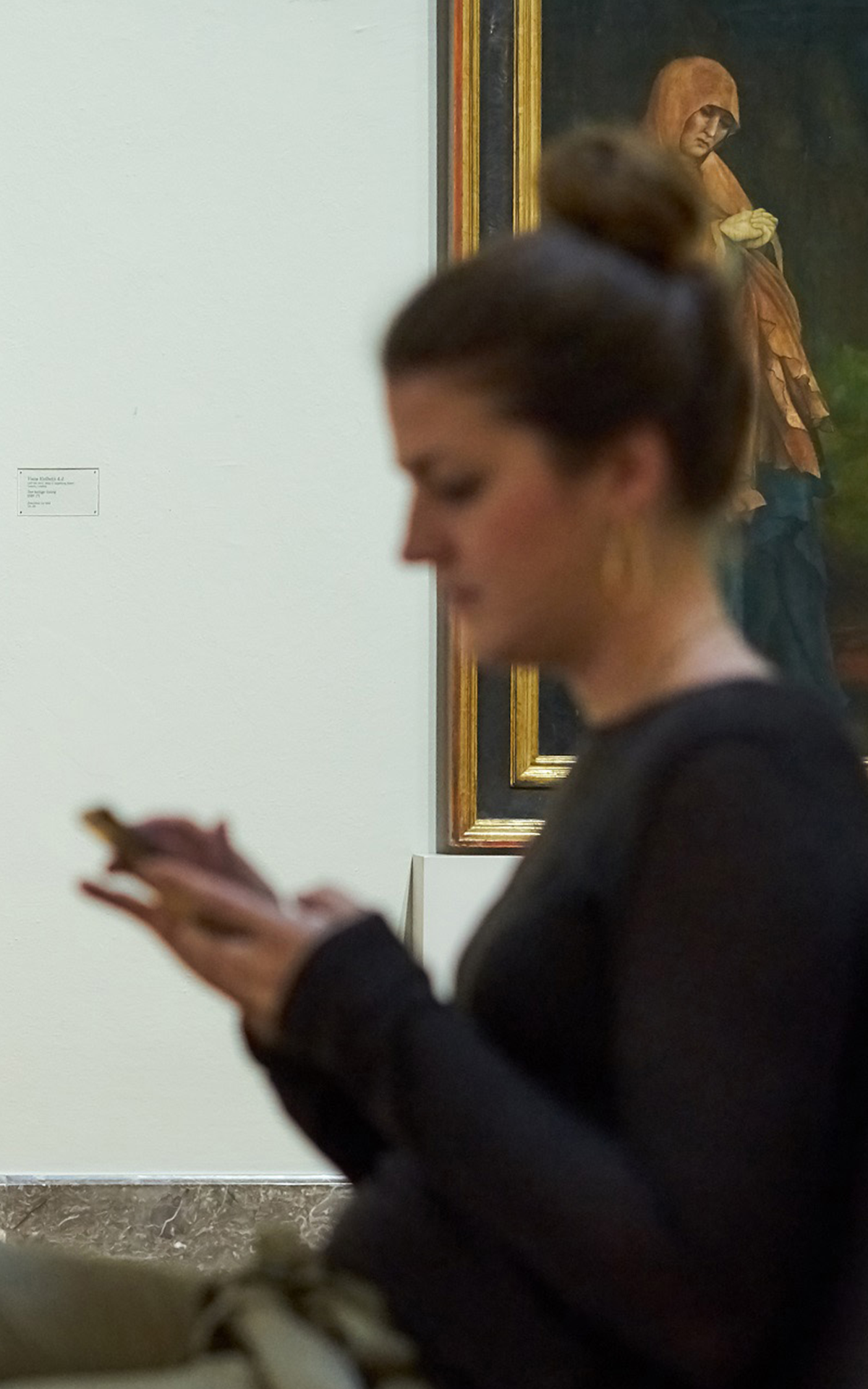 Junge Frau, die vor dem Werk eines Alten Meisters sitzend auf ihr Smartphone schaut