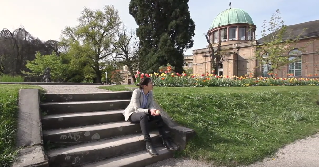 Mia Kukuk sitzt auf Stufen im botanischen Garten. Im Hintergrund ist die Orangerie zu erkennen.