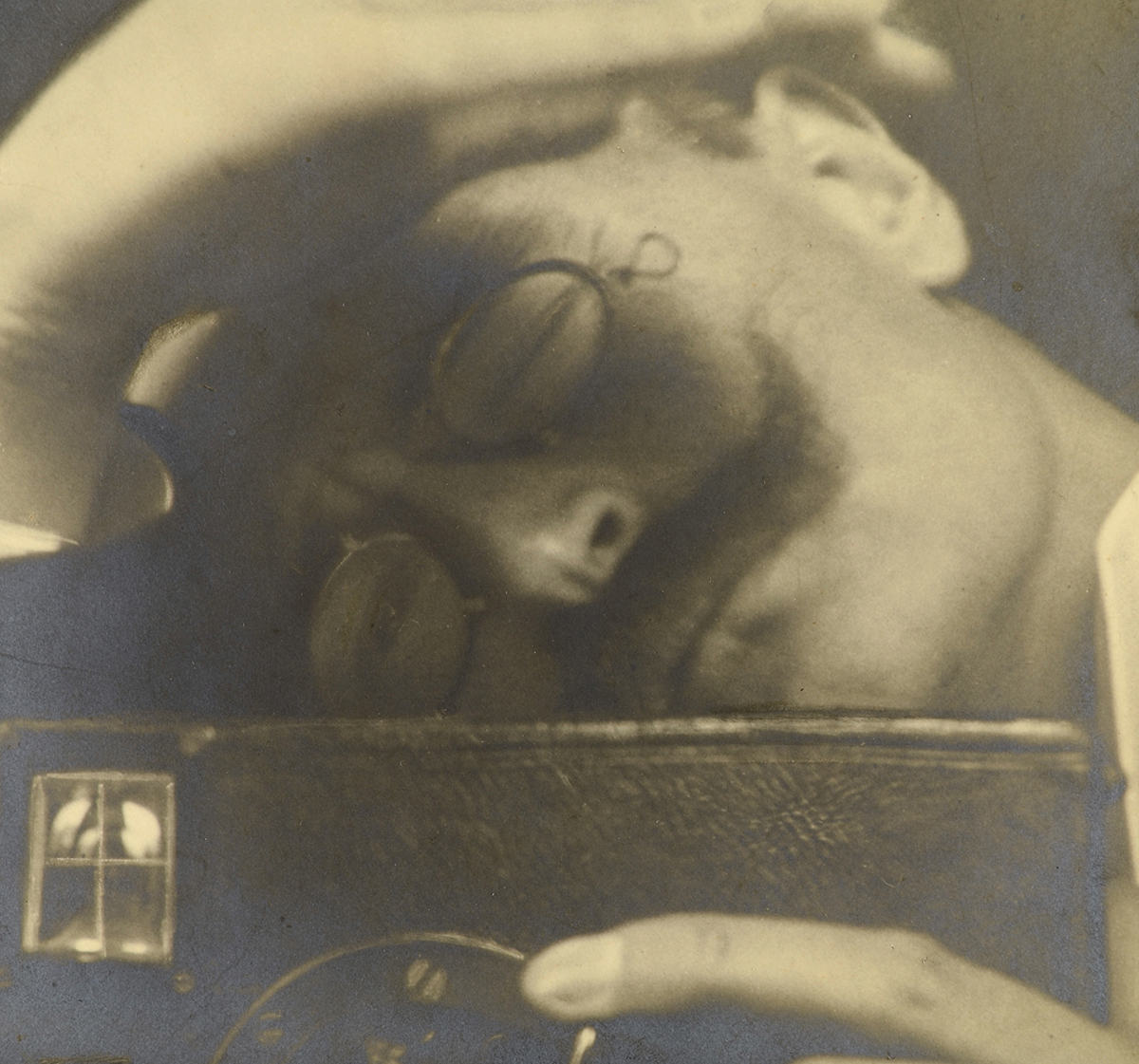Die Abbildung zeigt die Selbstfotografie Wilhelm Carl Friedrich Bandelow. Der Fotograf lehnt sich über seine Kamera, als benutze er es als Kissen und drückt ein Auge zu. Er trägt eine Brille und hat einen Schnäuzer.