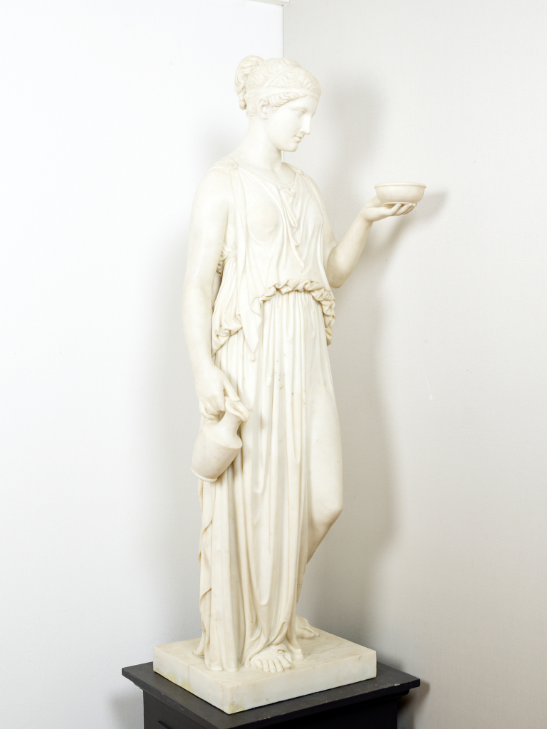 The classicist work of art by the artist and sculptor Bertel Thorvaldsen shows a woman in an antique robe. The sculpture is made of white marble and holds up a bowl.