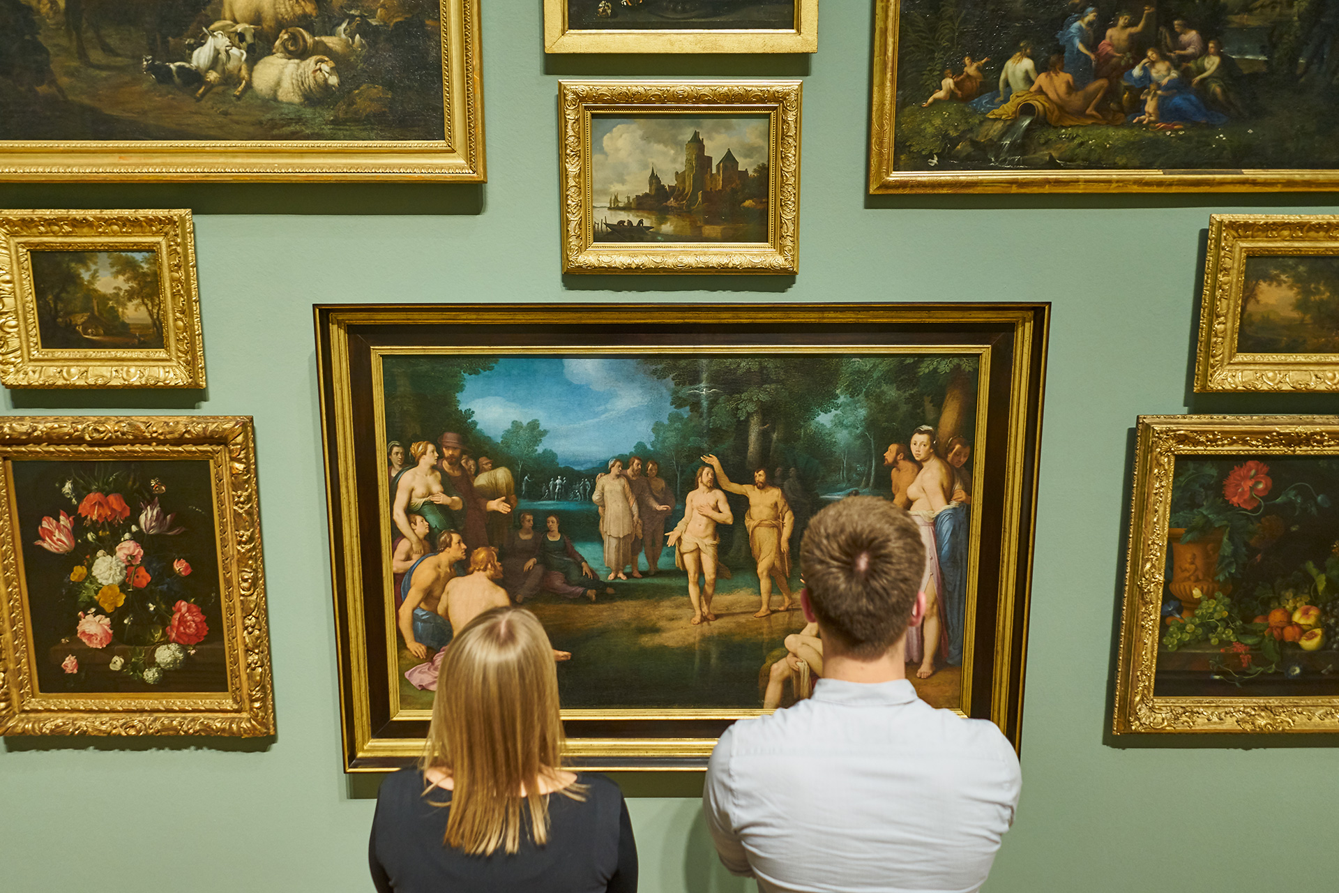 Eine Frau und ein Mann stehen vor einer Wand mit vielen Gemälden und sind in der Rückansicht erkennbar.