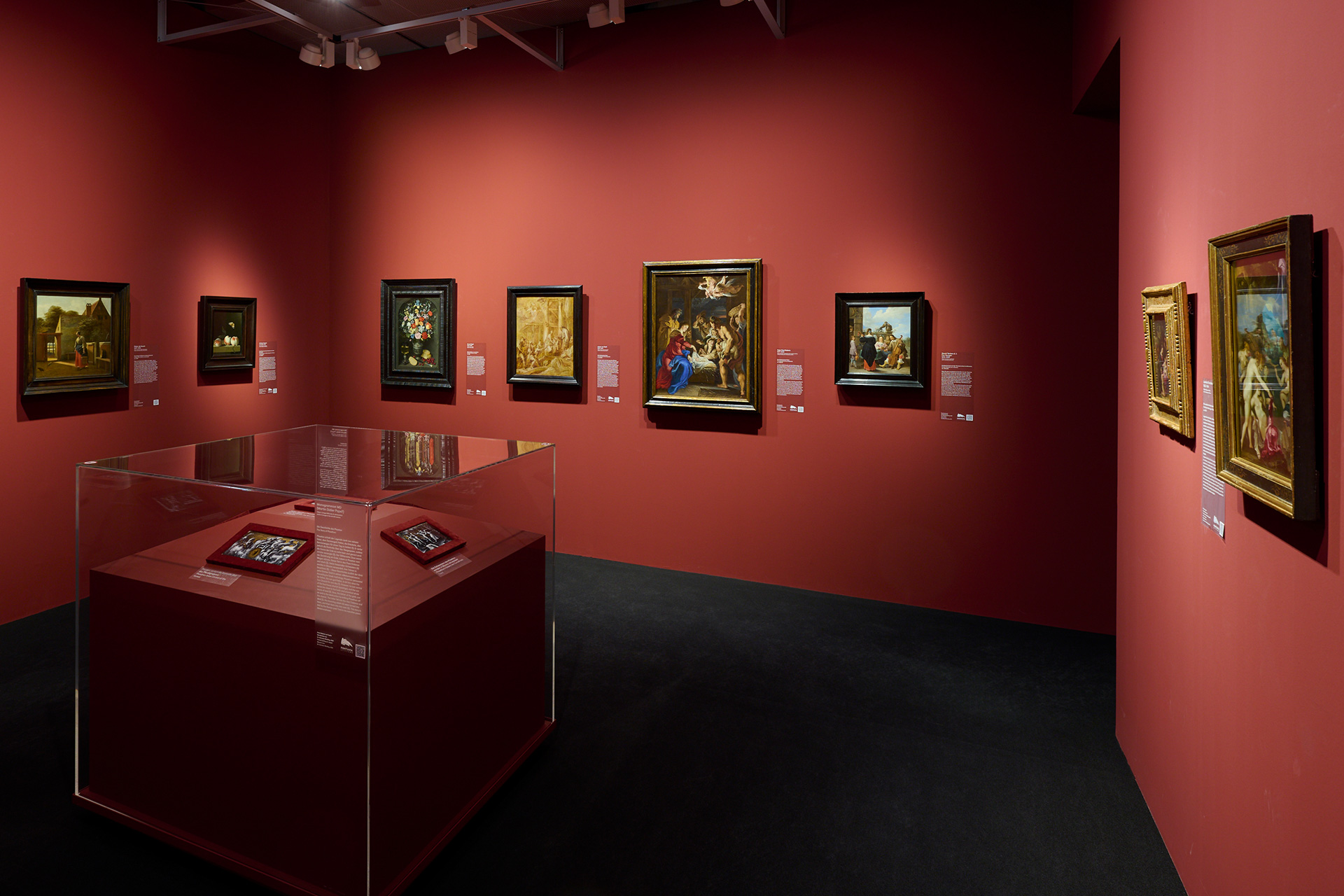 Ausstellungsansicht von einem roten Raum in dem einige Gemälde hängen. In der Mitte steht eine Vitrine.