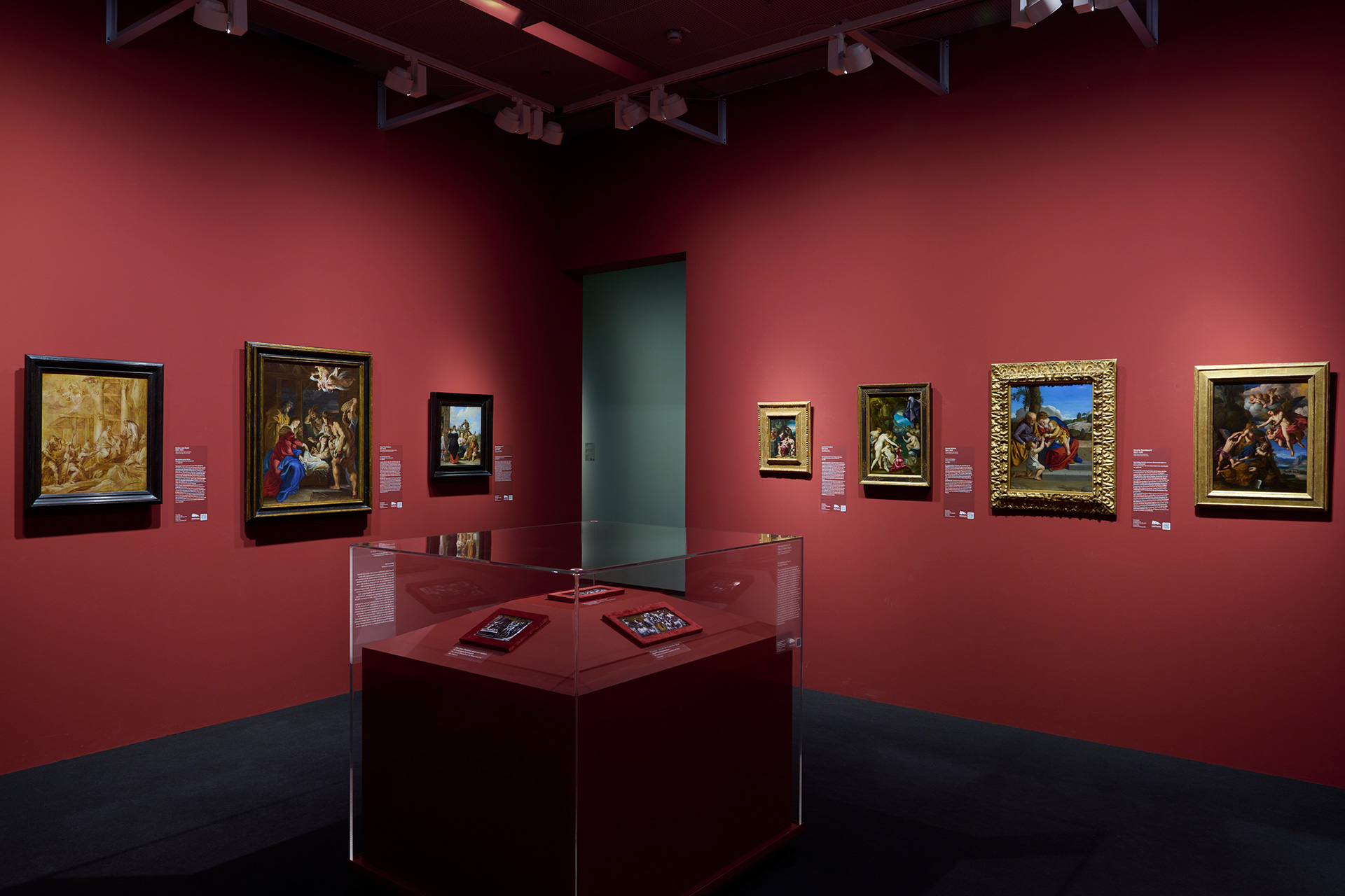 Auf der Ausstellungsansicht sieht man einen roten Raum, in dem verschiedene Gemälde hängen. In der Mitte des Raumes steht eine Vitrine.