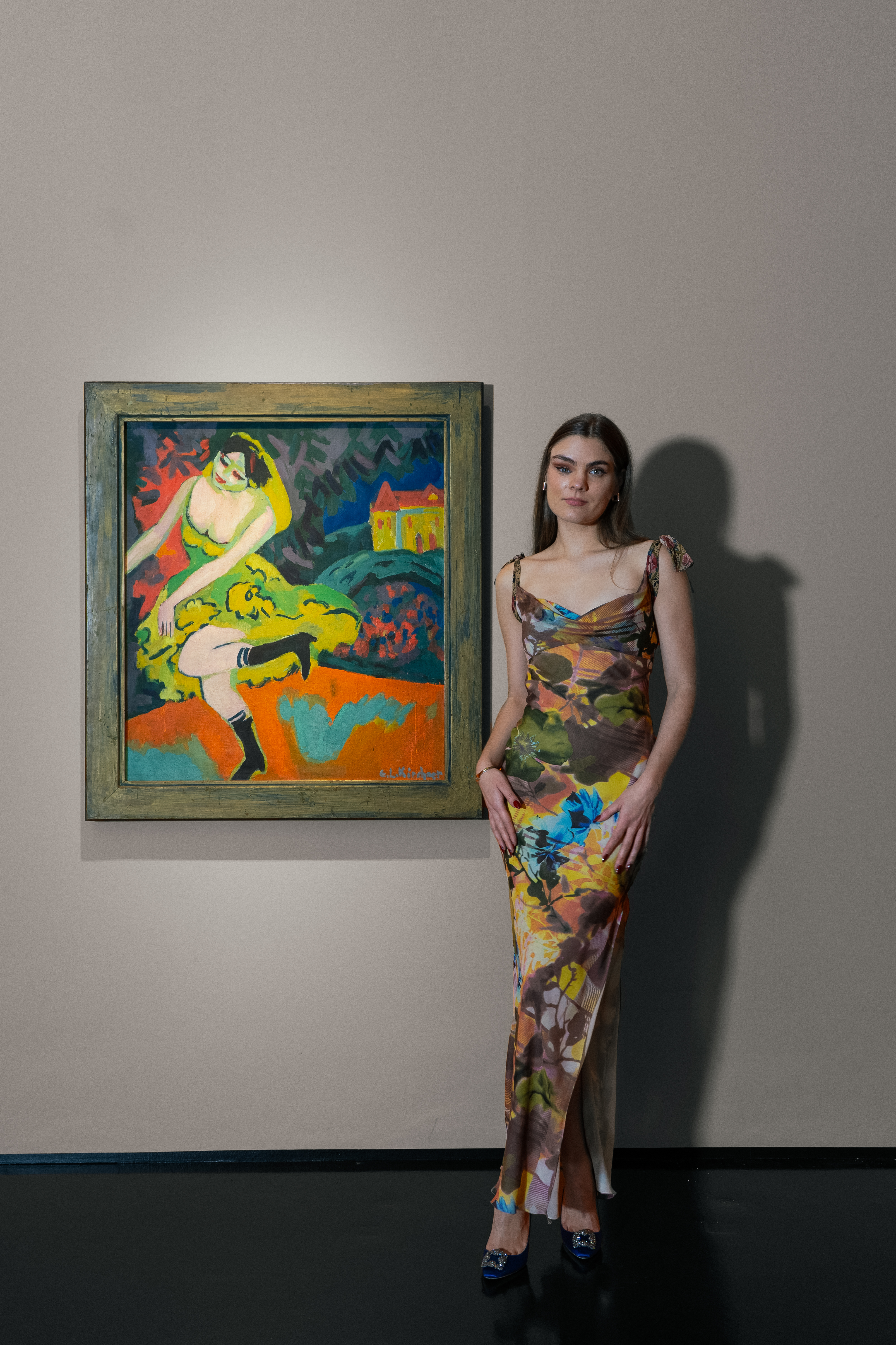 Eine Frau in einem bunten enganliegendem Kleid neben dem farbigen Werk von Kirchner, auf dem man eine tanzende Frau sieht.