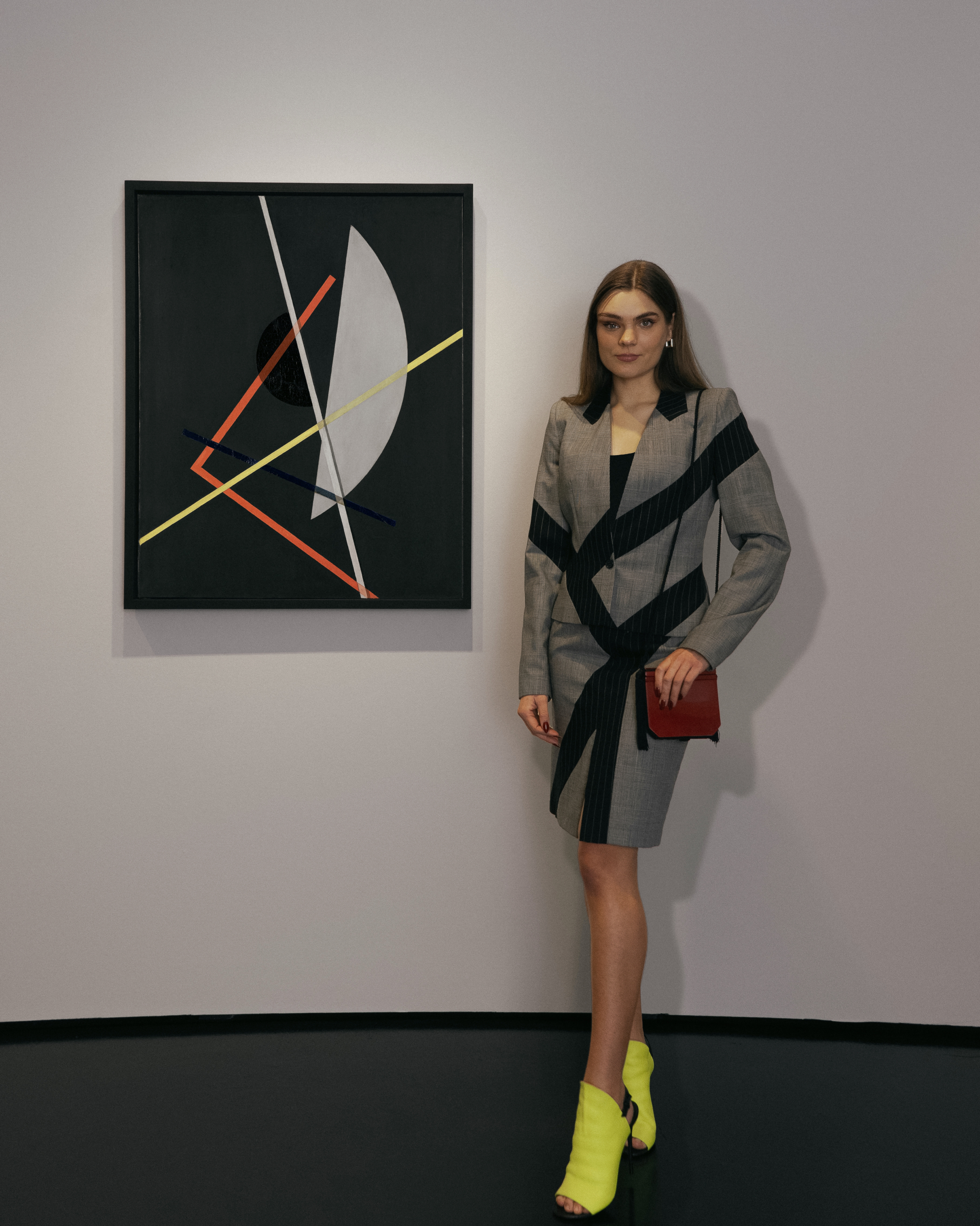 Eine Frau mit einem grauen Kostüm mit schwarzen Streifen und neongelben Schuhen neben dem Werk von Moholy-Nagy, auf dem man verschiedenen geometrische Formen, wie Kreise und Linien sieht.
