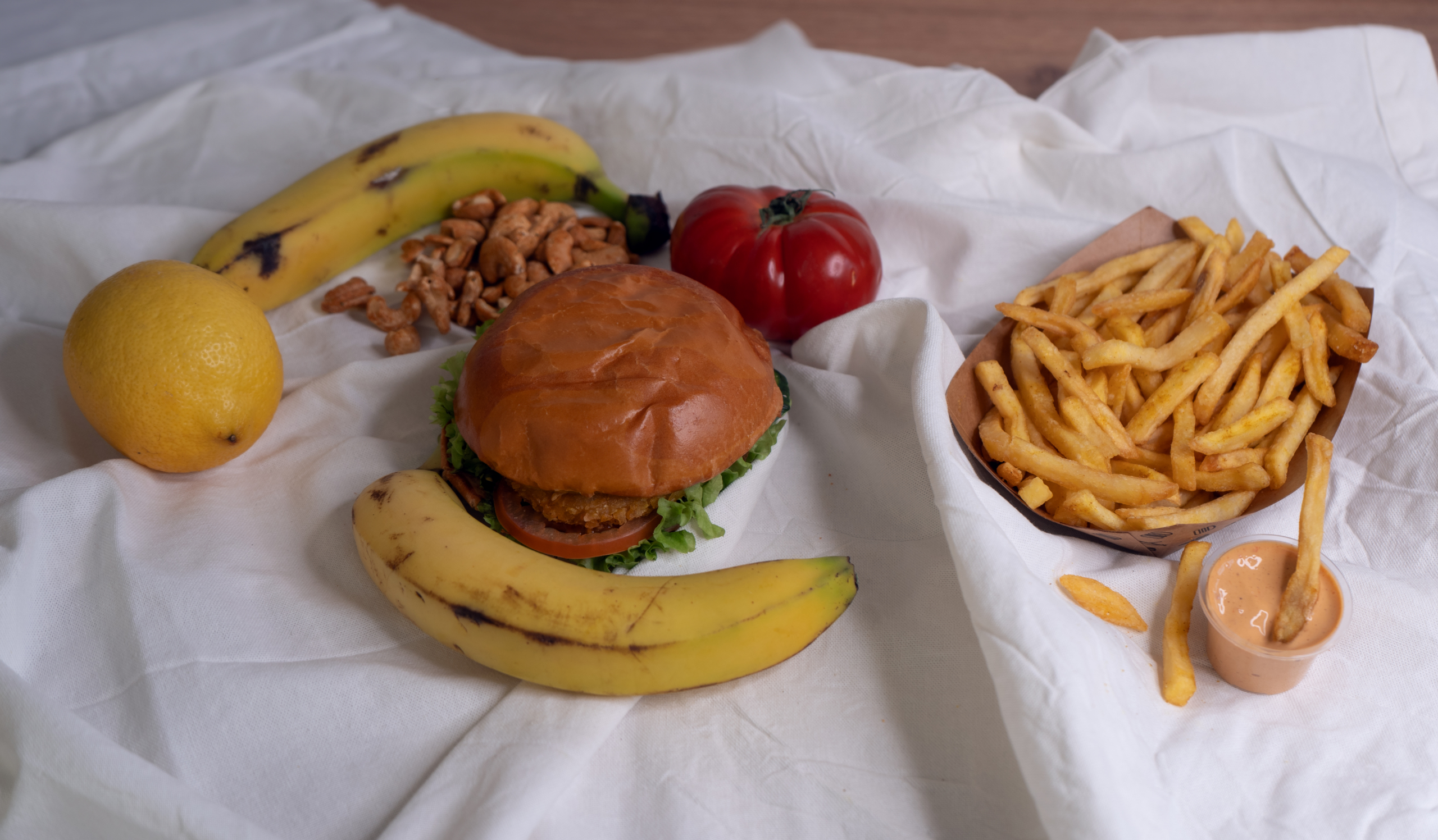 Ein Stillleben auf dem zwei Bananen, eine Zitrone, eine Tomate, Cashewkerne, ein Burger und Pommes sind.