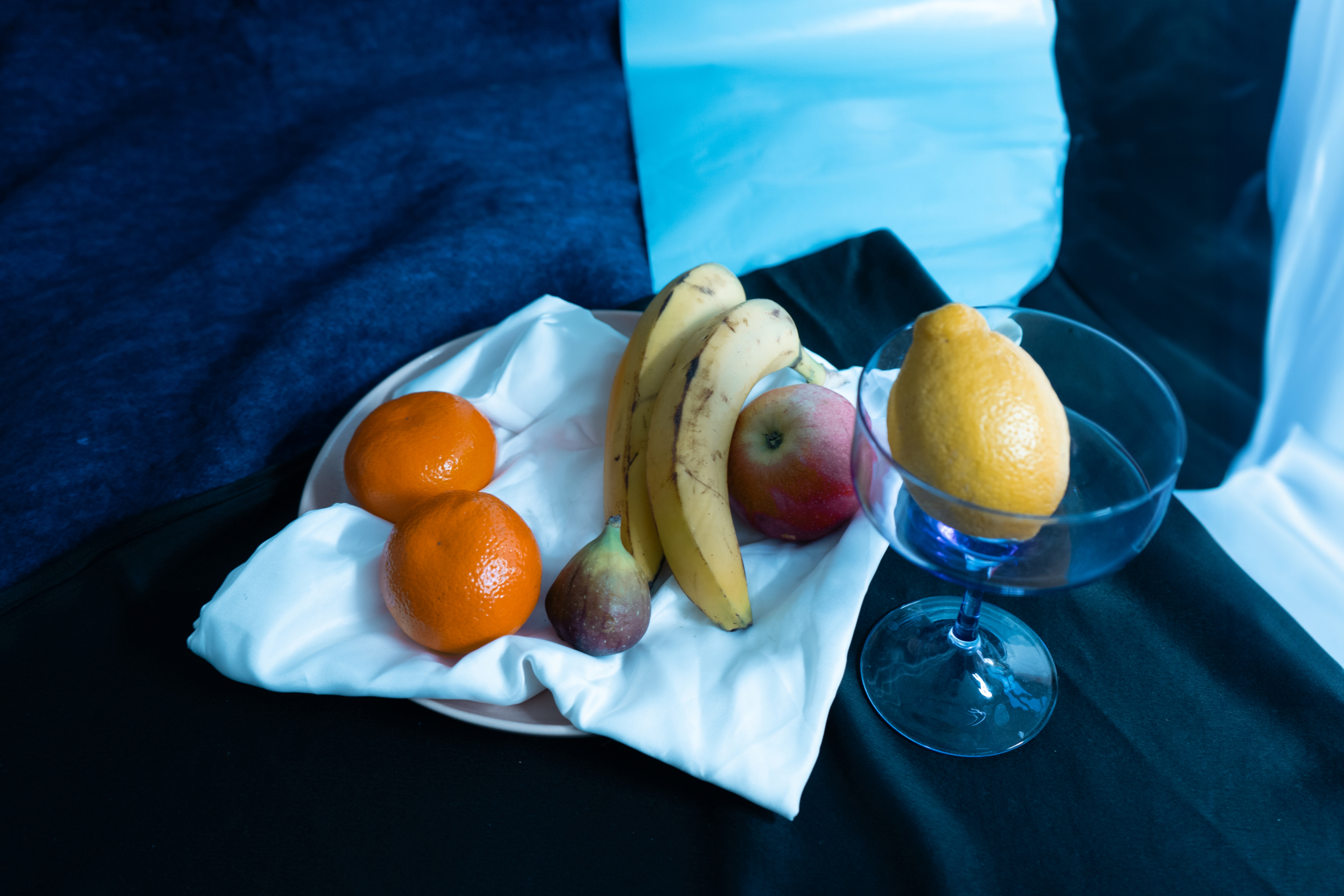 Ein Arrangement aus einer Zitronen, zwei Bananen, einer Feiger, zwei Mandarinen und einem Apfel vor einem blauen Hintergrund.