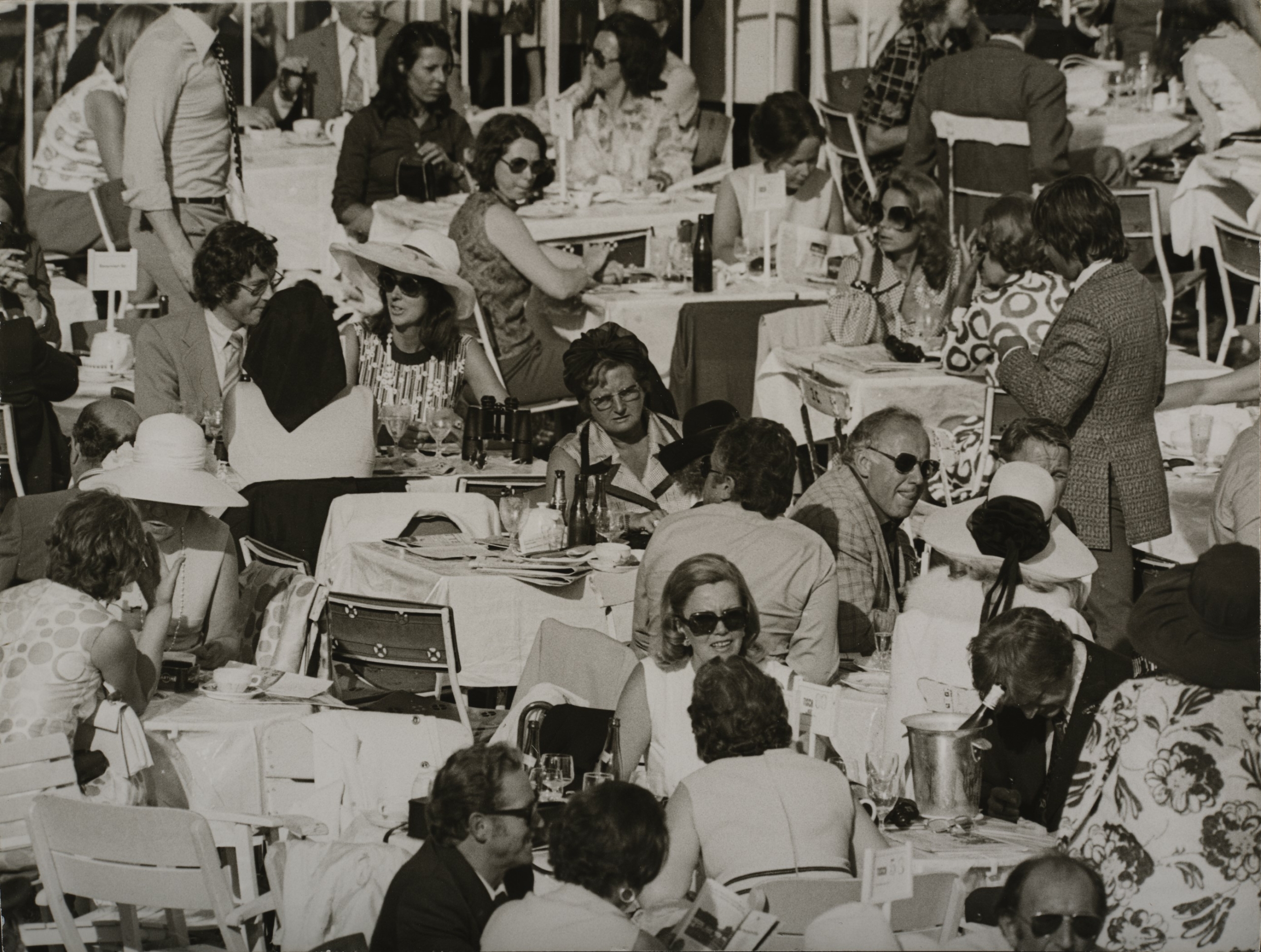 Die Fotografie zeigt Personen, die im Freien an Cafe-Tischen sitzen, sich unterhalten, essen und trinken.
