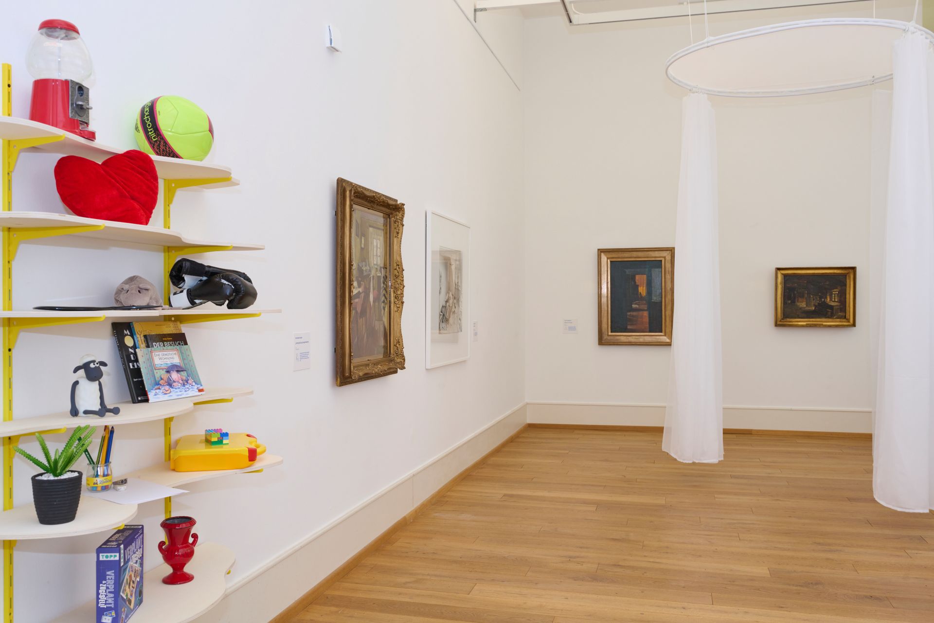 Blick in den Ausstellungsraum der Jungen Kunsthalle. Bilder und ein mit Spielzeug befülltes Regal hängen an der Wand.