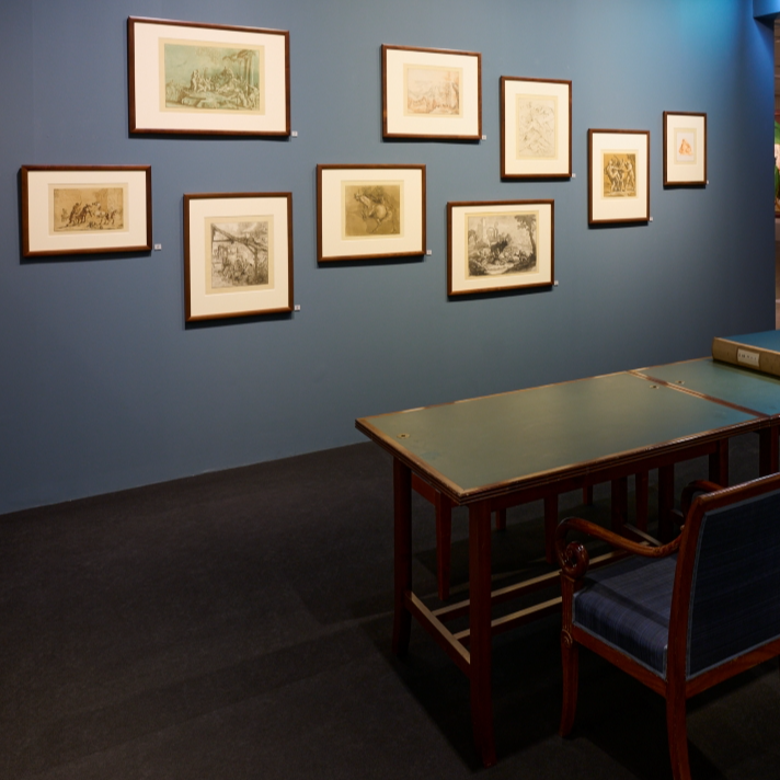 Impression der Ausstellung über Druckgrafiken des Ehepaars Prestel. In der Raummitte steht ein Holzschreibtisch mit Stuhl, dahinter hängen gerahmte Grafiken an einer blauen Wand.
