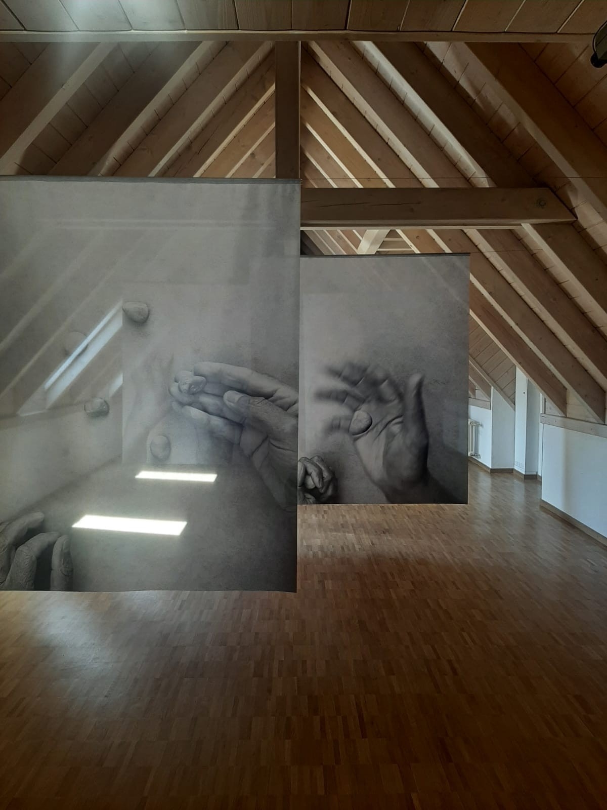 Blick in die Ausstellung "Crossing Traces". Der Ausstellungsraum ist in einem Dachgeschoss, die Holdbalken des Dachgiebels laufen spitz zu. Von Querbalken hängen die großfrmatigen schwarz-weiß Bilder, die Hände zeigen, die spielerisch mit Steinen hantieren.