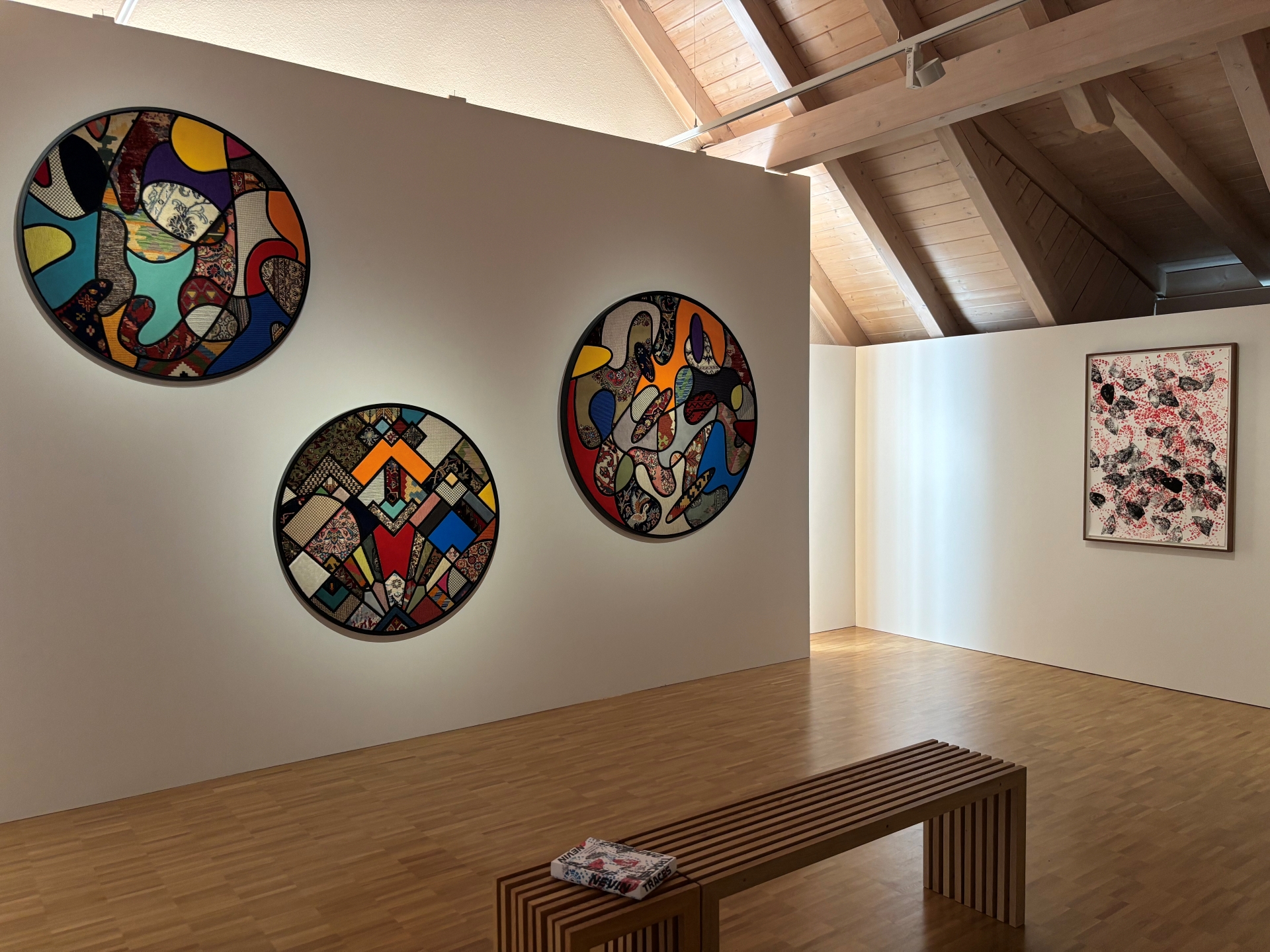 Blick in den Ausstellungsraum von "Crossing Traces" mit Holzvertäfelter Decke und Holzboden. An einer weißen Ausstellungswand hängen drei kreisförmige Textilarbeiten in bunten Farben.