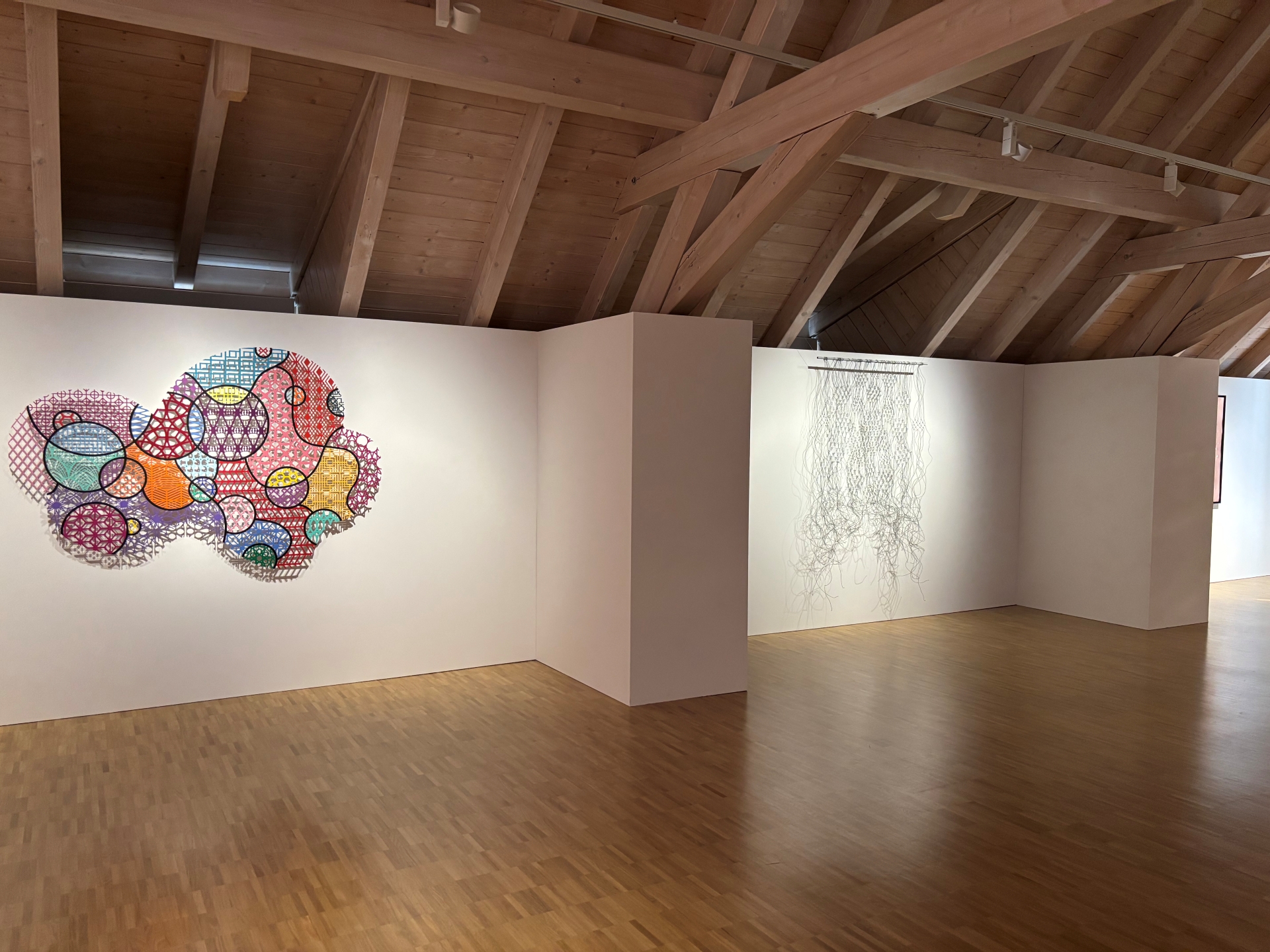 Blick in den Ausstellungsraum von "Crossing Traces" mit holzvertäfelter Decke und Holzboden. An dem weißen Ausstellungswänden hängt ein aus Draht geknüpftes Makramee sowie ein buntes Wandobjekt in wolkenform.