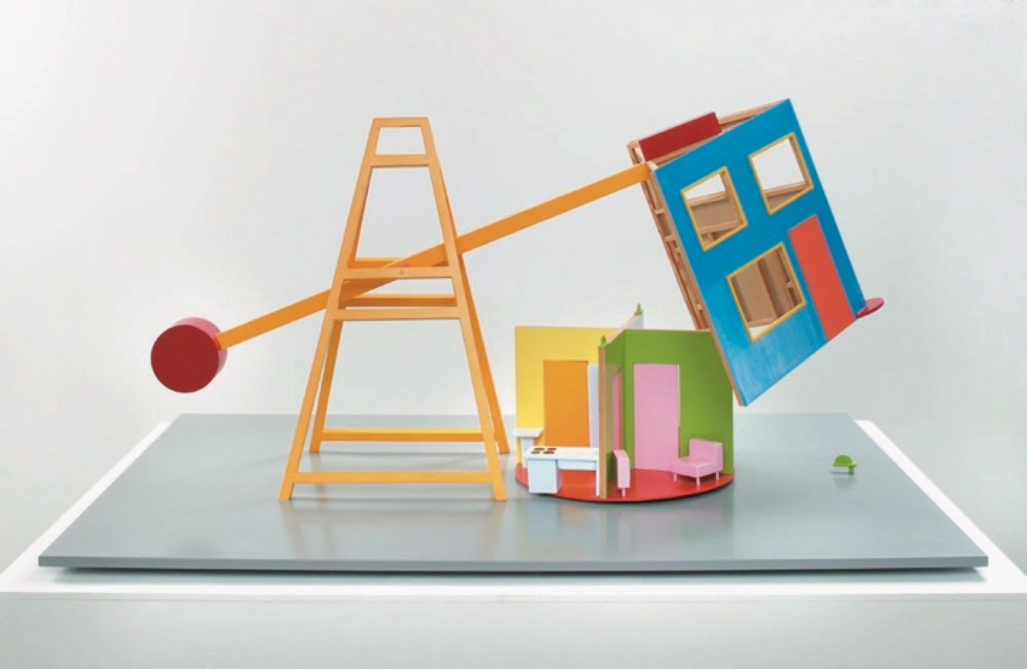 Das bunte Architekturmodell besteht aus einem Gerüst mit einem Pendel, an dessen Ende ein Dach befestigt ist. Schwingt das Pendel, so weht das Dach über dem nebenstehenden kleinen Haus.