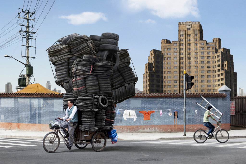 A man rides a bicycle, towing a trailer loaded with car tires tied together into a gigantic tower. In the background, a massive high-rise complex looms.