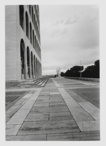 Die Schwarzweißfotografie zeigt den langen Vorplatz des Palazzo della Civiltà Italiana. Auf der linken Seite ist seine Fassade zu sehen. Am Ende des Vorplatzes steht eine Statue.