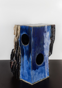 Eine ca. 50 cm hohe Skulptur steht auf dem Boden. Sie besteht aus unterschiedlichen zusammengesetzten Formen. Ihre Oberfläche ist teilweise glatt oder rau.