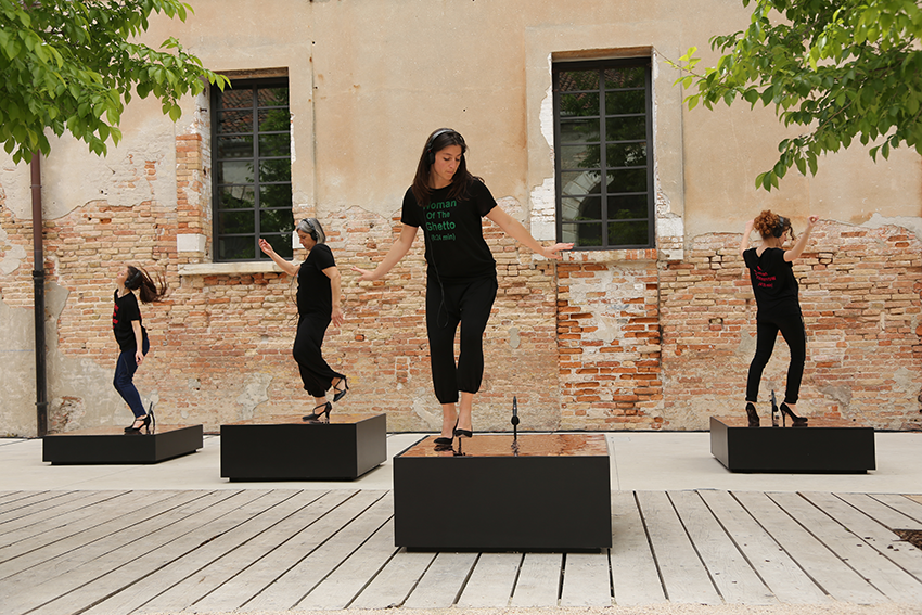 Foto einer Performance: Vier Frauen tanzen auf einzelnen Podesten vor einer Mauer mit Fenstern, sie alle tragen schwarze Kleidung und Schuhe.