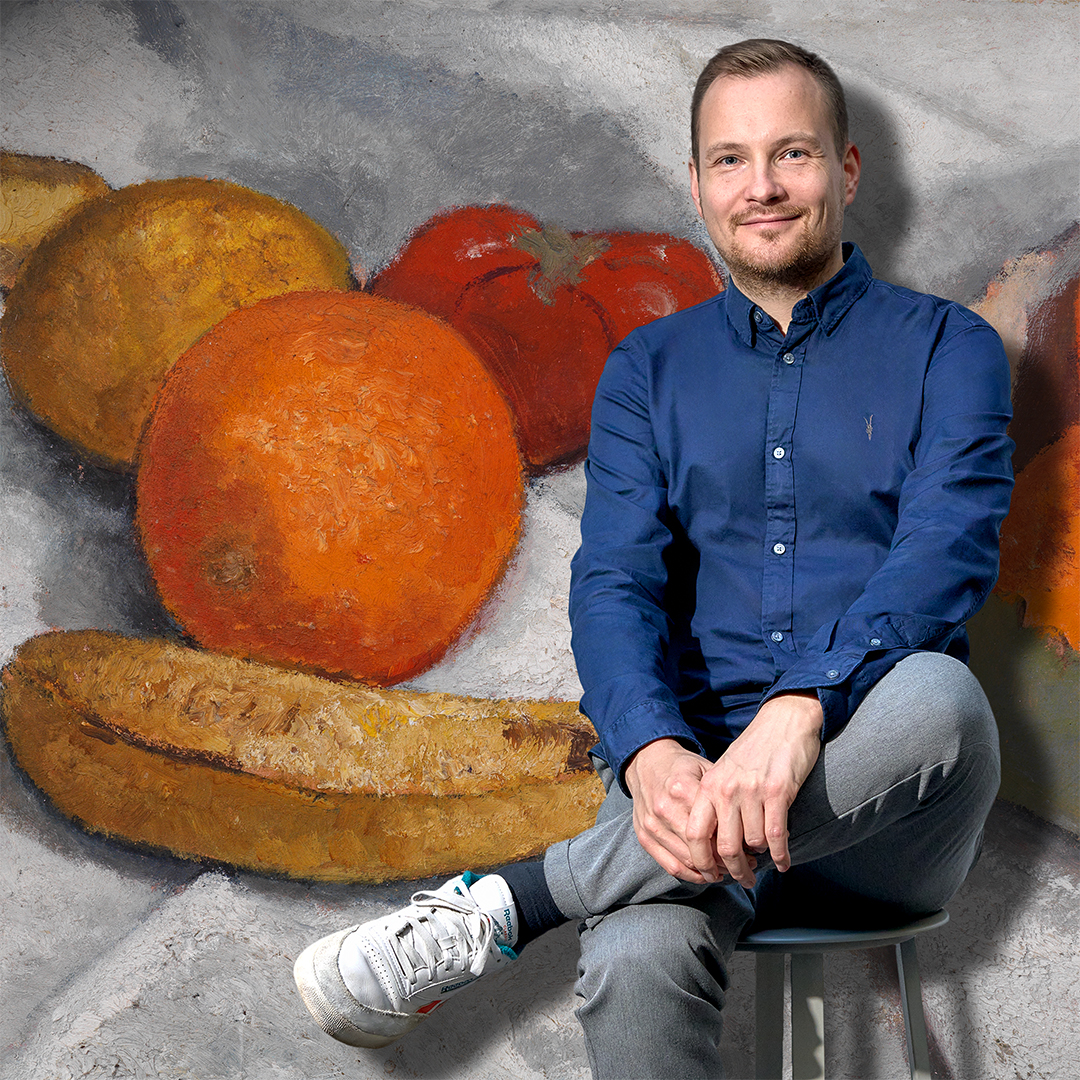 Foto des Podcasters Jakob Schwerdtfeger, hinter ihm ist das Stilleben mit Orangen, Bananen, Zitronen und Tomate der Künstlerin Modersohn-Becker zu sehen.