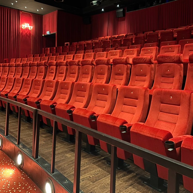 Blick in ein Kino auf leere Kinoreihen mit roten Samtsesseln, rotem Teppichboden und Samtvorhängen.