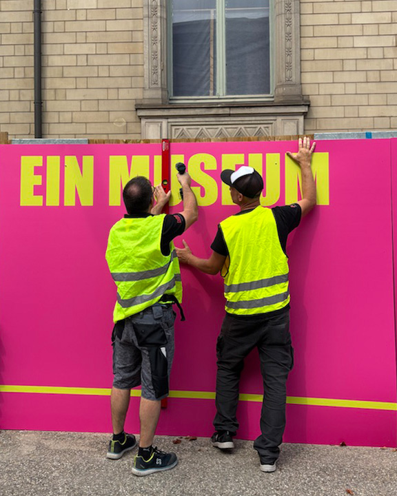 Zwei Männer in gelben Warnwesten bringen ein Zaunbanner vor dem Hauptgebäude der Kunsthalle an. Sie nutzen Wasserwaage und Bohrer. Das Banner ist pink und hat eine gelbe Aufschrift auf der steht: Ein Museum.