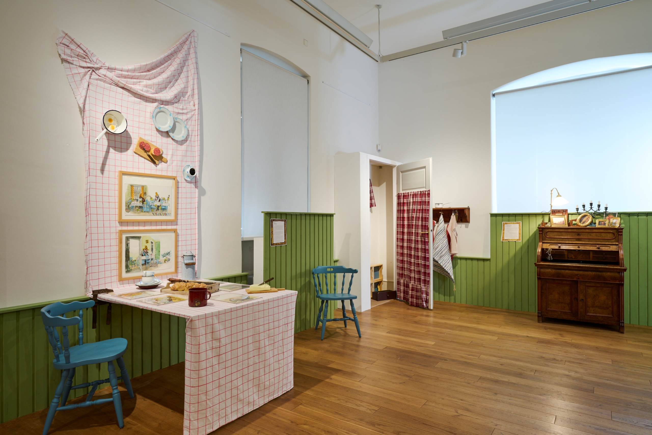 Blick in den Ausstellungsraum der Jungen Kunsthalle. Die Wände sind halbhoch mit grüner Vertäfelung ausgestattet. Im Raum steht ein Frühstückstisch mit Karodecke. An den Wänden hängen Zeichnungen.