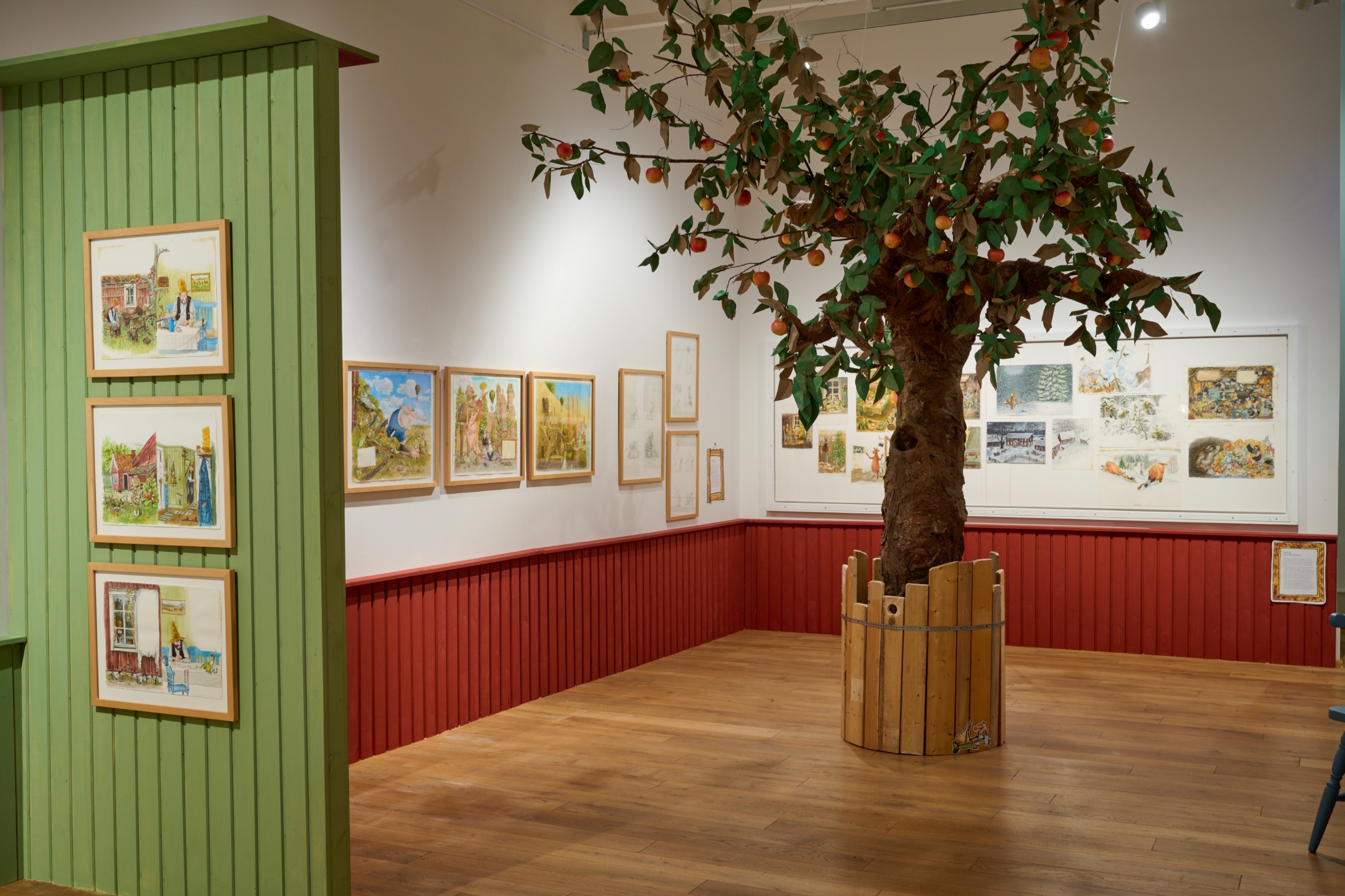 Blick in den Ausstellungsraum der Jungen Kunsthalle. Die Wände sind halbhoch mit roter Vertäfelung ausgestattet. An den Wänden hängen gerahmte Ilustrationen. In der Mitte des Raumes steht ein Apfelplastikbaum.