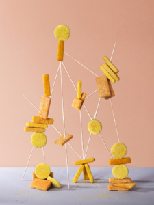 Verschiedene Fischstäbchen gestapelt und auf Holzspieße gesteckt, bilden abstrakte geometrische Strukturen vor einem schlichten Hintergrund.