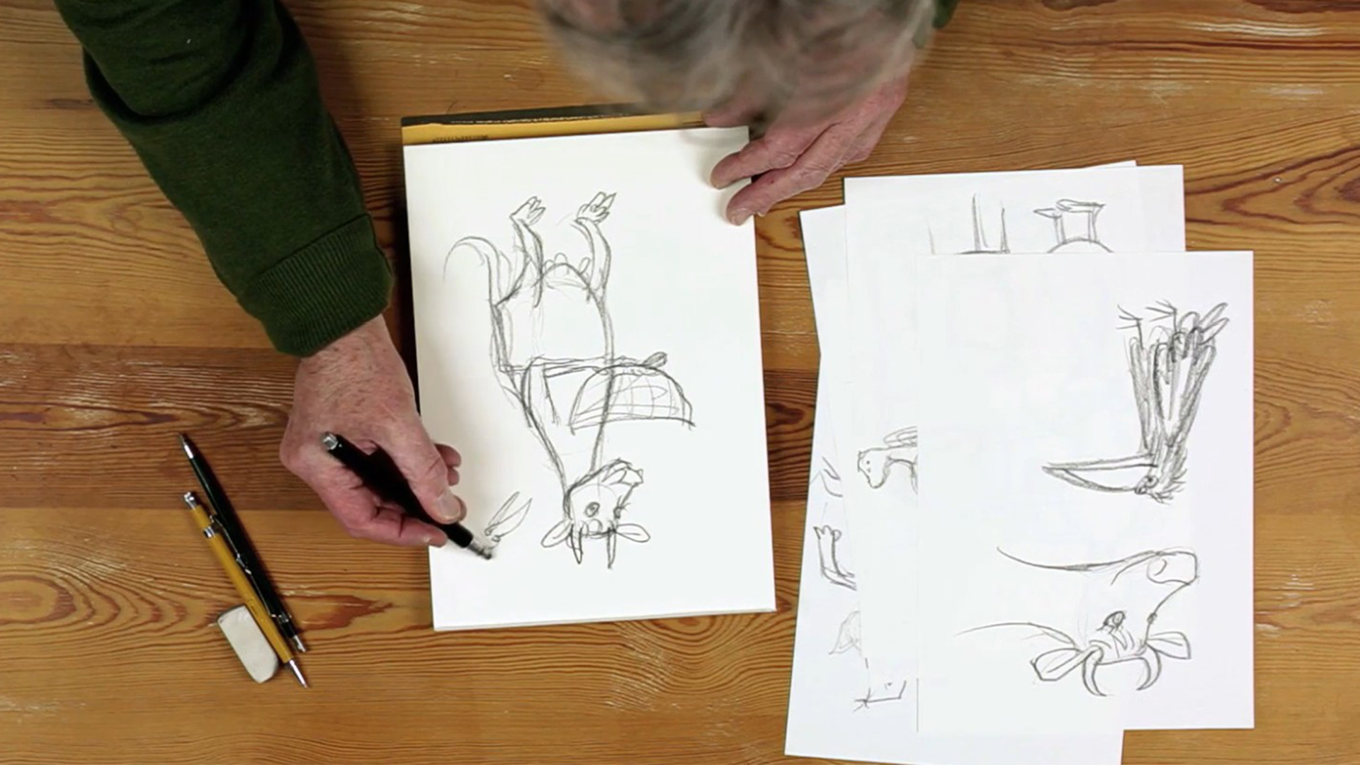 Aus der Vogelperspektive auf einen Tisch an dem man zwei Händen sieht, die auf einem Skizzenblatt einen Kuh skizzieren.