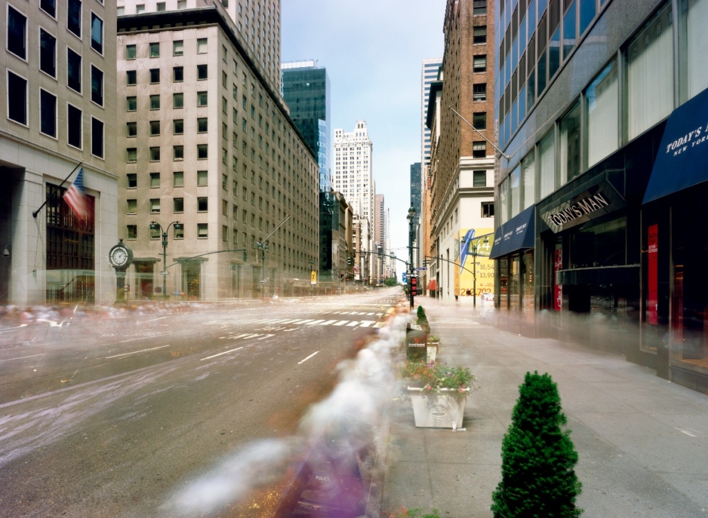 Eine breite Straße ist rechts und links von Hochhäusern gesäumt. Der Nebel über der Straße zeichnet die Spuren der Menschen, die während der Aufnahme dort waren, nach.