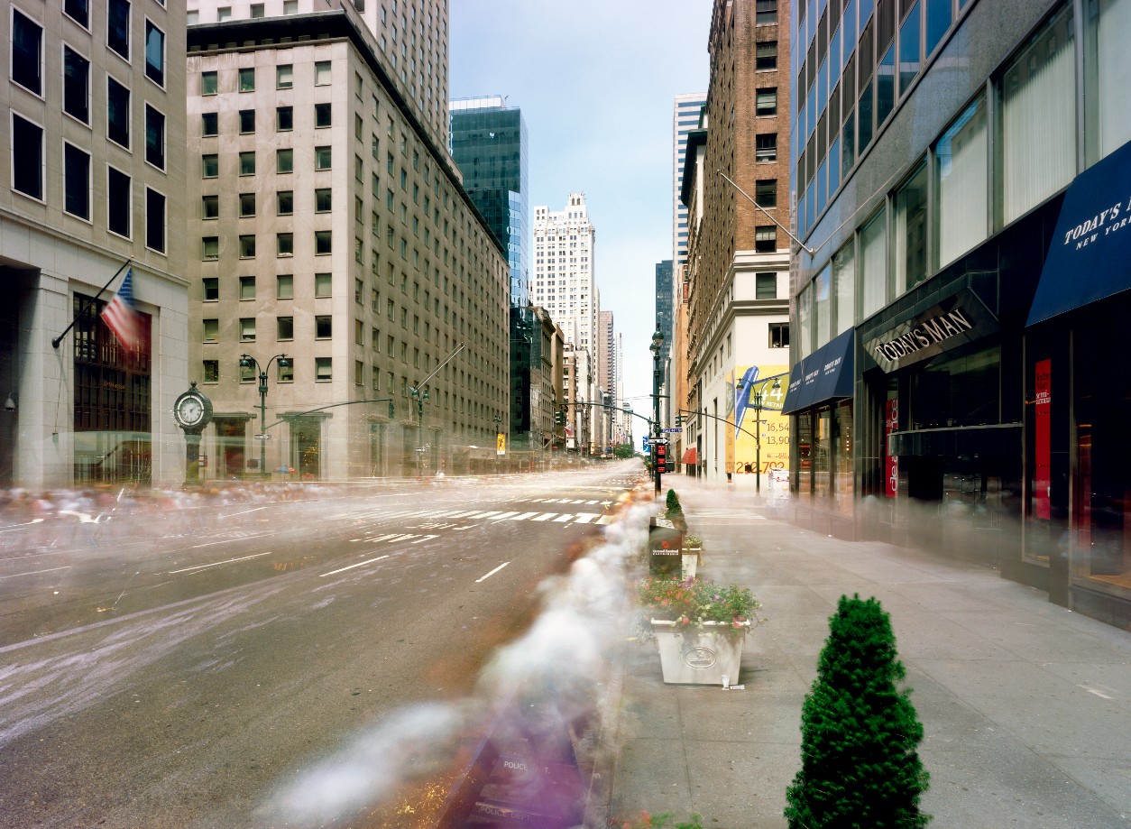 Eine breite Straße ist rechts und links von Hochhäusern gesäumt. Der Nebel über der Straße zeichnet die Spuren der Menschen, die während der Aufnahme dort waren, nach.