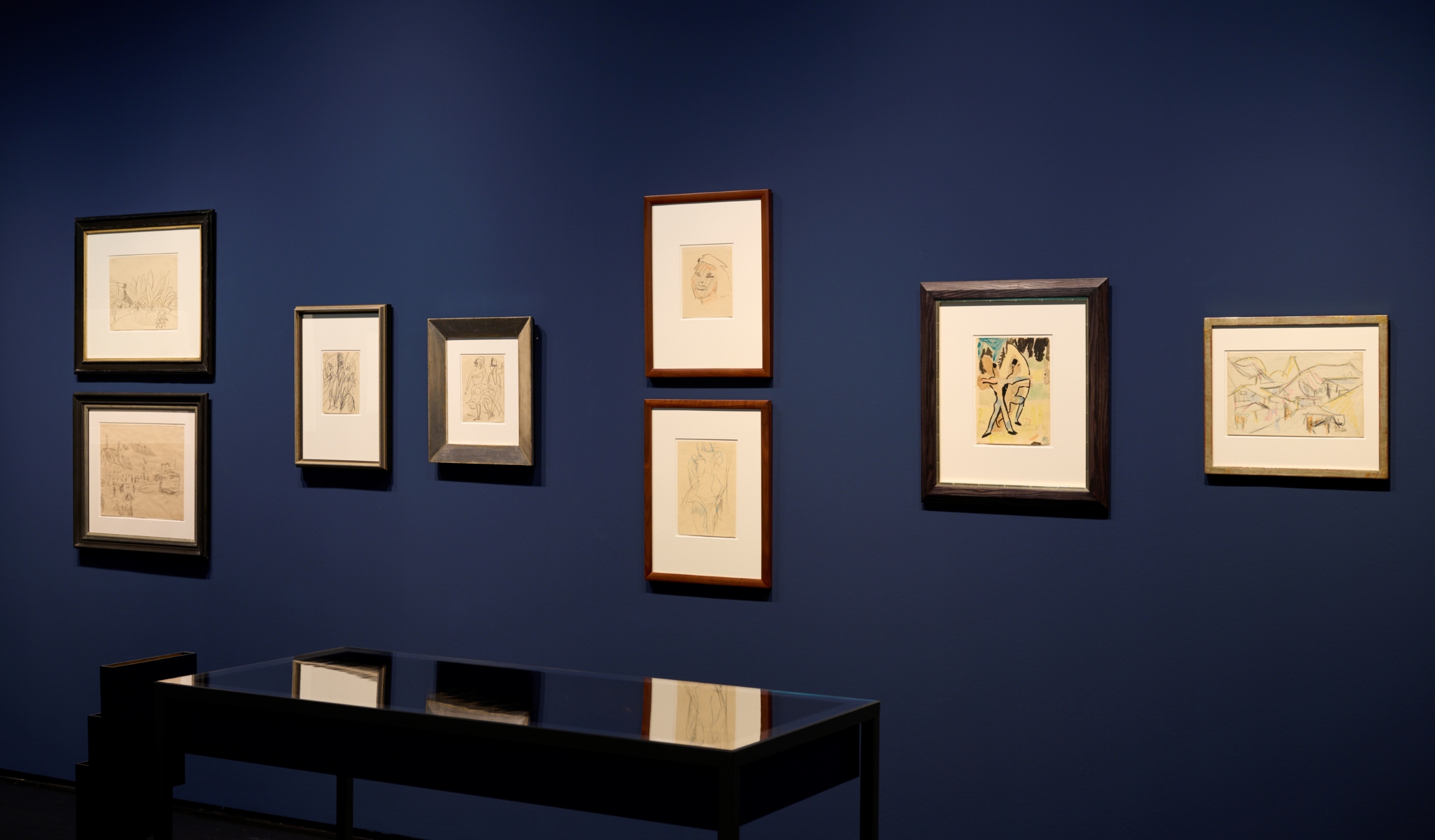 Blick in den Ausstellungsraum der Kunsthalle mit dunkelblauen Wänden. An den Wänden hängen viele kleine gerahmte Zeichnungen auf weißem und gelben Papier. Davor steht ein Schaukasten, der die Bilder an der Wand durch das Glas reflektiert.