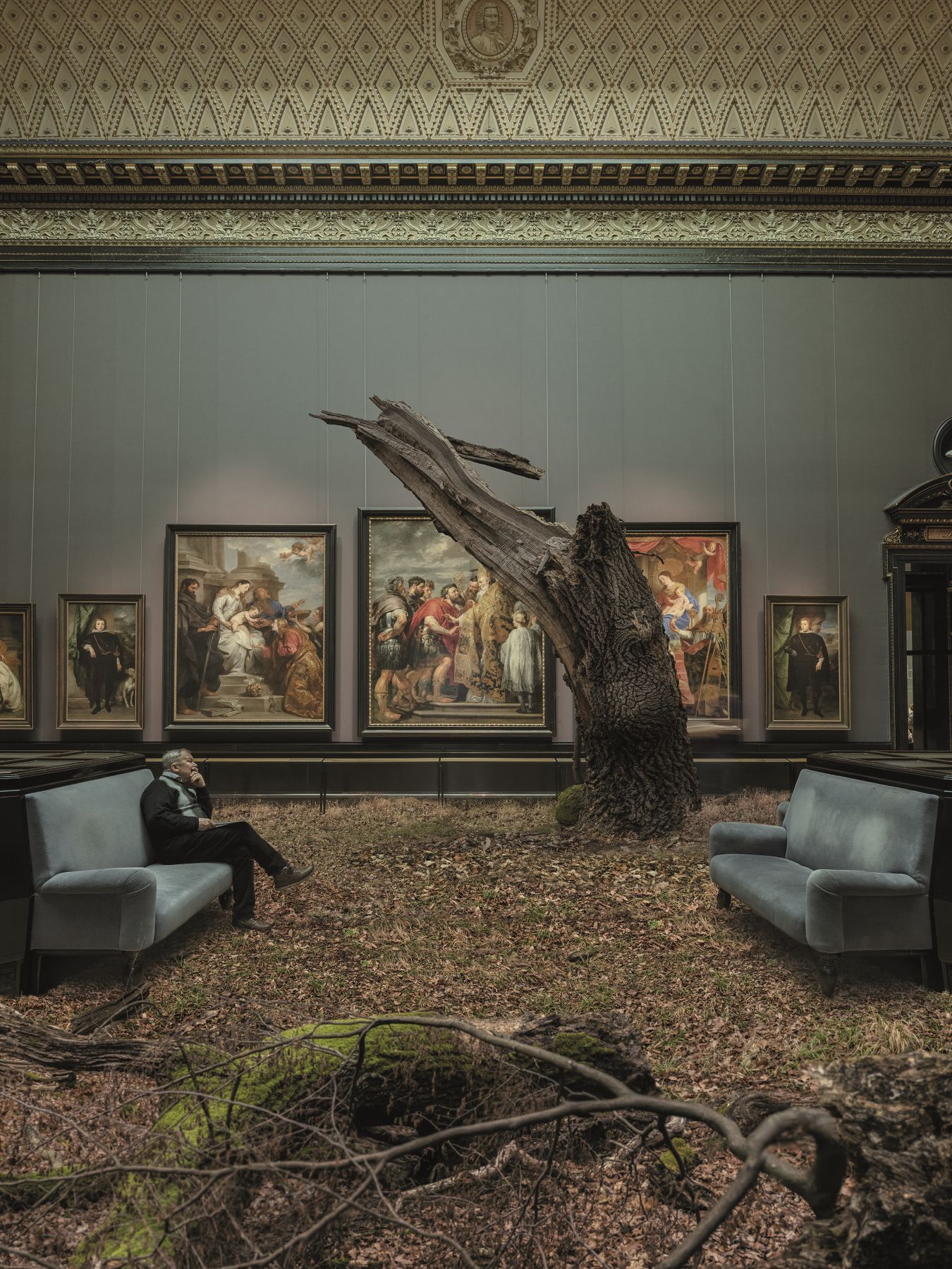 Zu sehen ist der Raum eines Museums. Der Boden ist vollständig mit braunen Blättern bedeckt, ein abgebrochener Baum ragt in der Mitte des Raumes aus dem Boden. Auf einer Bank in der Raummitte sitzt ein Mann, der das Gesicht auf seine Hand stützt. An den Wänden hängen großformatige Kunstwerke.