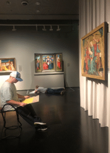 Blick in die Ausstellung mit christlichen Kunstwerken, ein auf einem Museumshocker sitzender junger Erwachsener arbeitet.